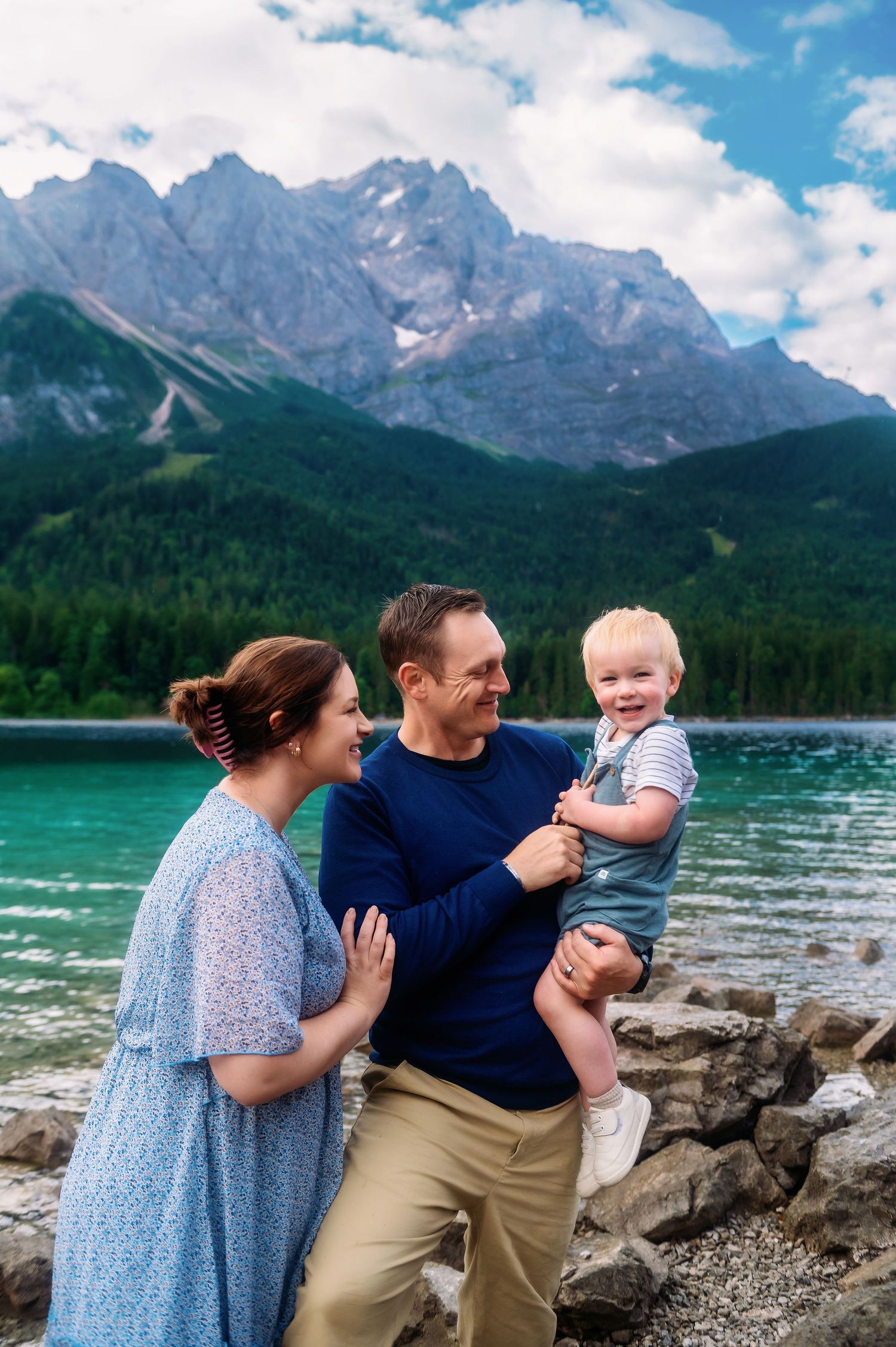 Family of three posing by a turquoise alpine lake with dramatic mountain peaks and lush forest in the background.