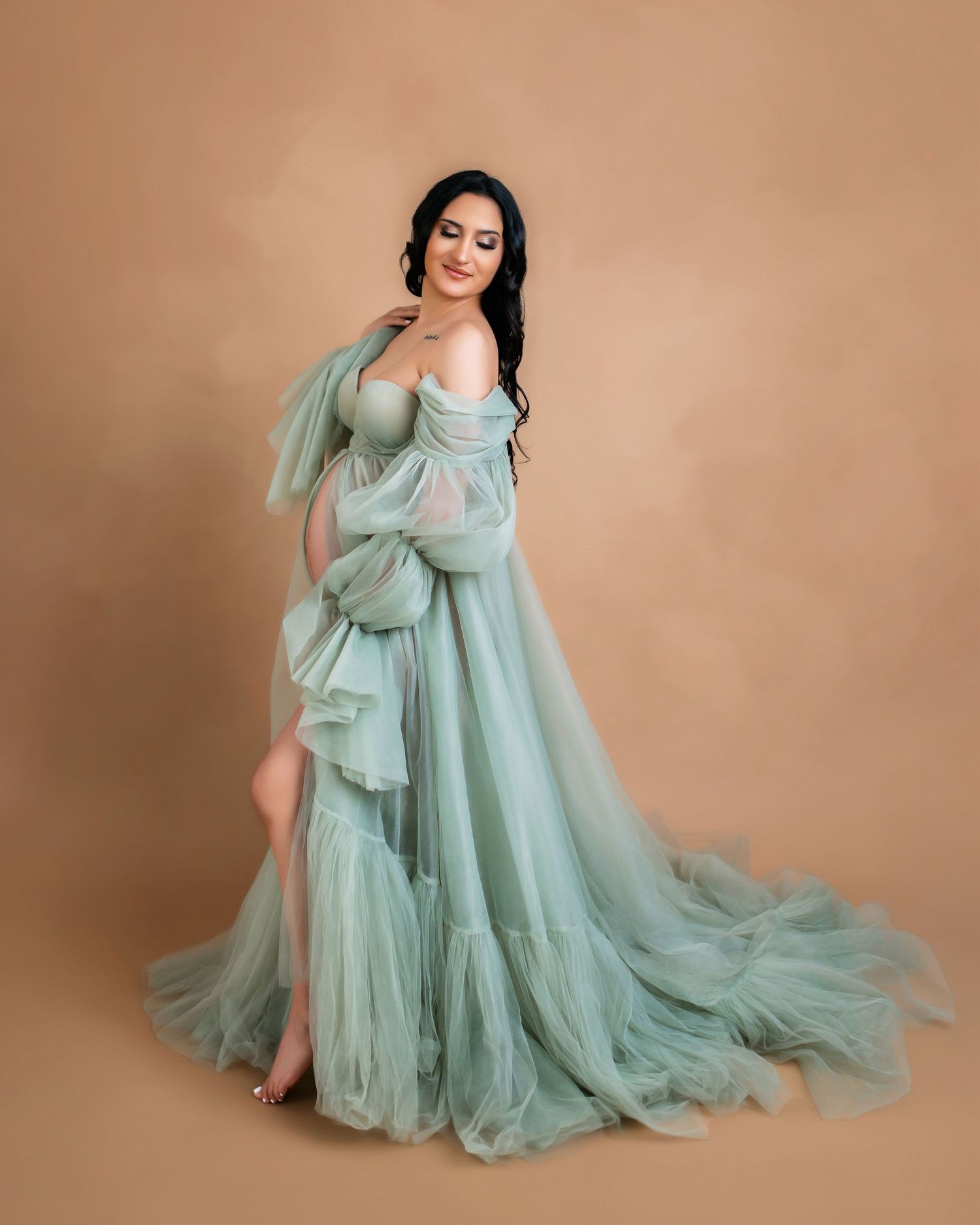Woman in elegant sage green tulle maternity gown posing gracefully against a warm beige studio background.