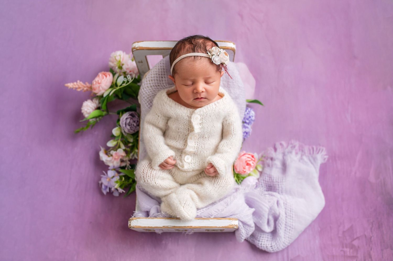 Sleeping newborn baby wearing white lace outfit lying on purple backdrop with delicate floral decorations.