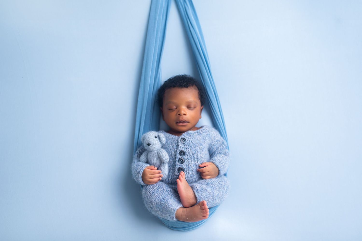 Sleeping newborn in blue knit outfit nestled in suspended fabric hammock.