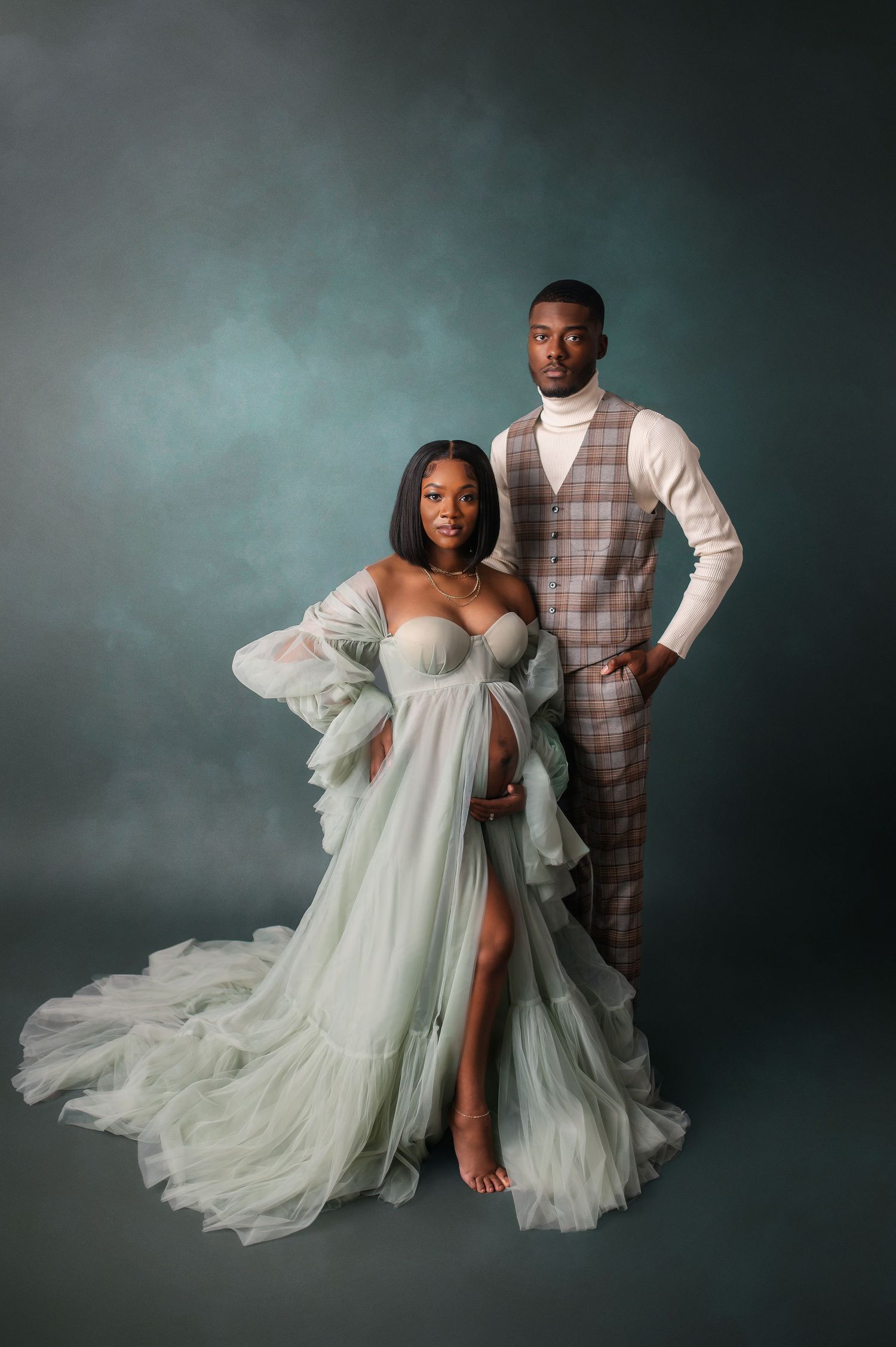 Romantic maternity photoshoot with couple in formal attire against moody teal background.