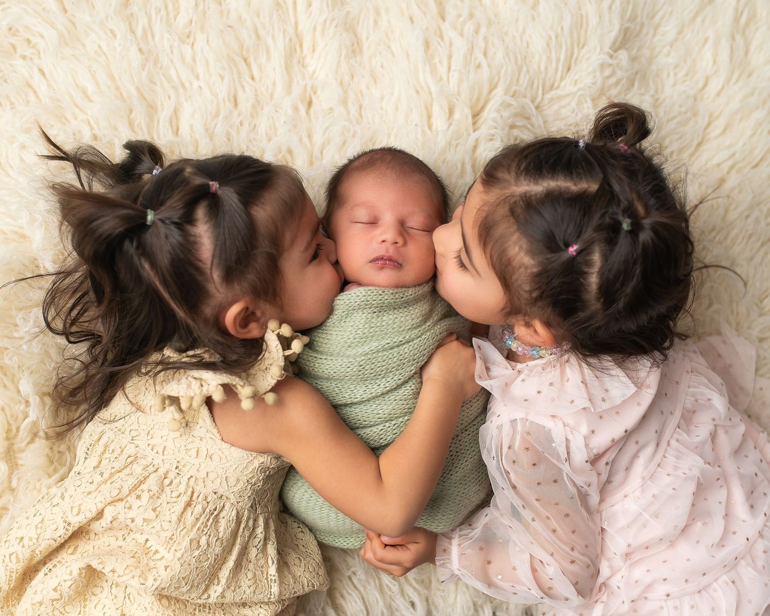 Three children lying on cream-colored fabric with a sleeping newborn in a mint green wrap in the center.