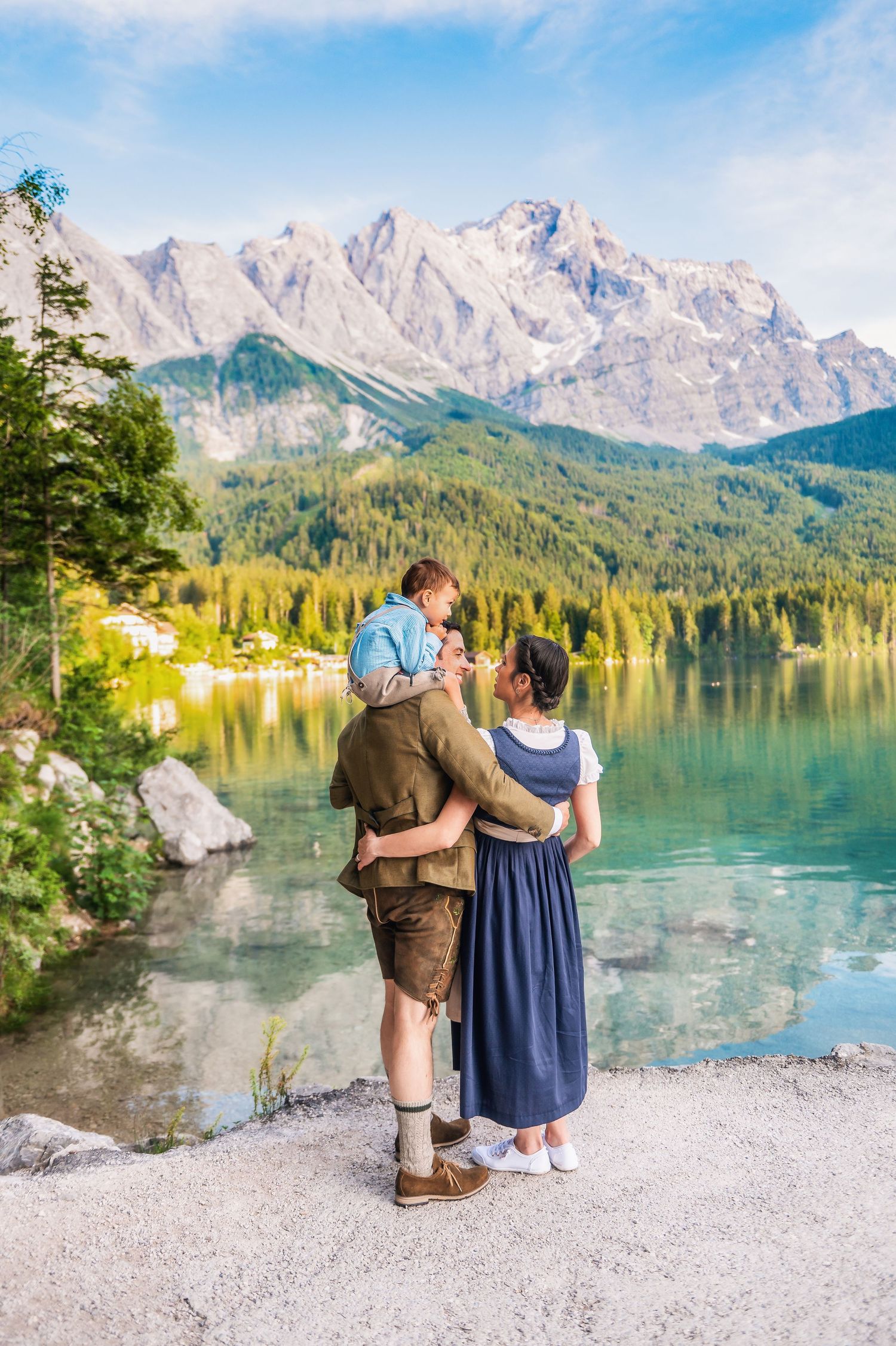 A picturesque Alpine lake reflects snow-capped mountains while two people in traditional Bavarian dress sit on the shoreline.