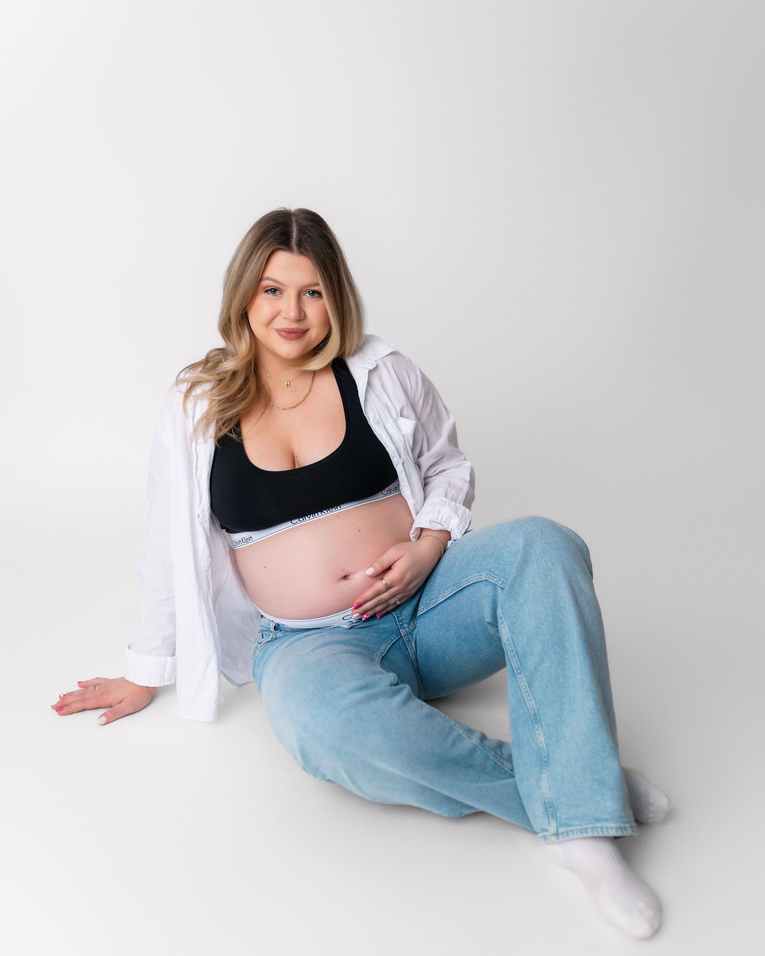 Pregnant woman in black top and white shirt poses sitting on floor during studio photoshoot.