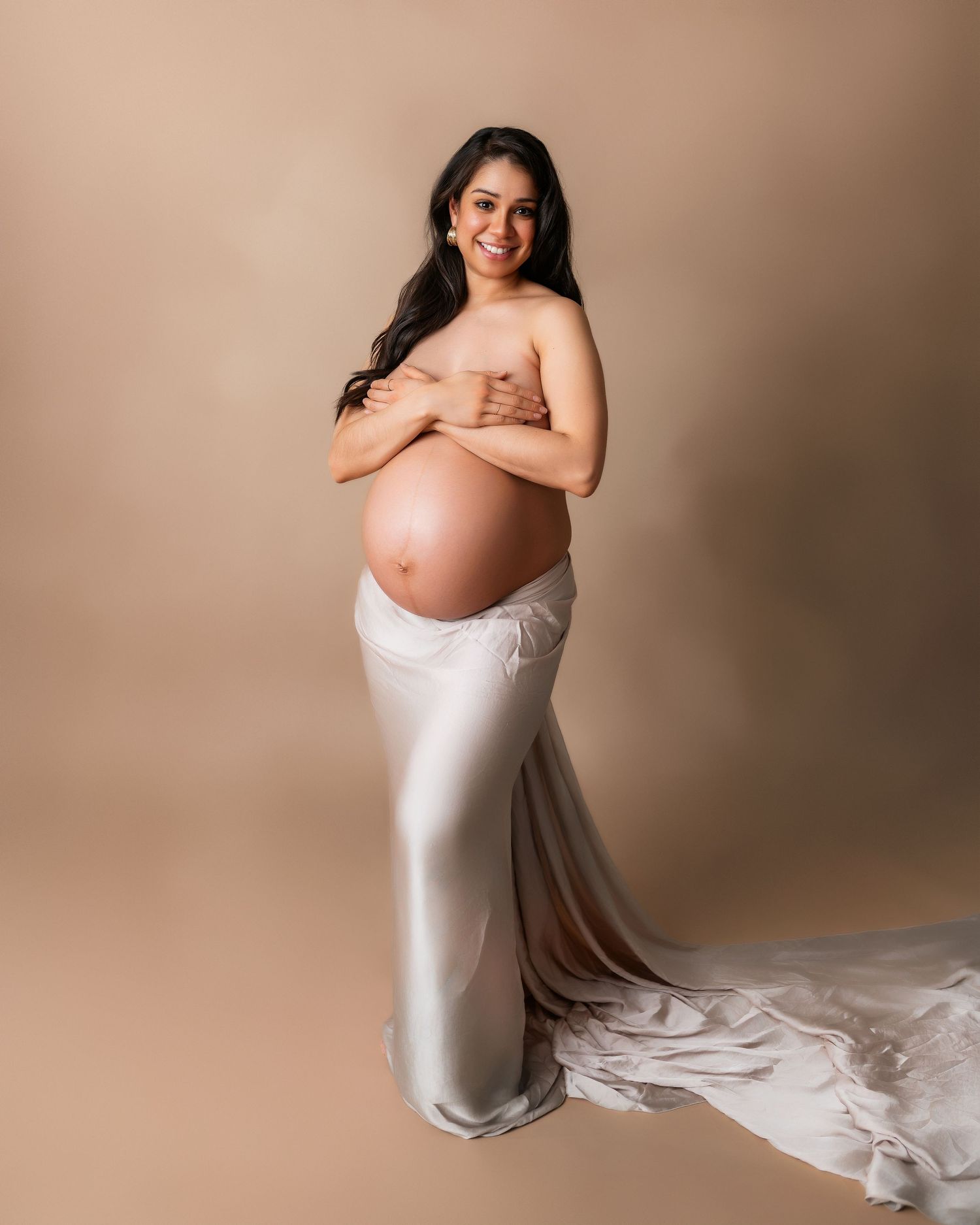 Elegant maternity portrait with woman in white silk gown against neutral background.