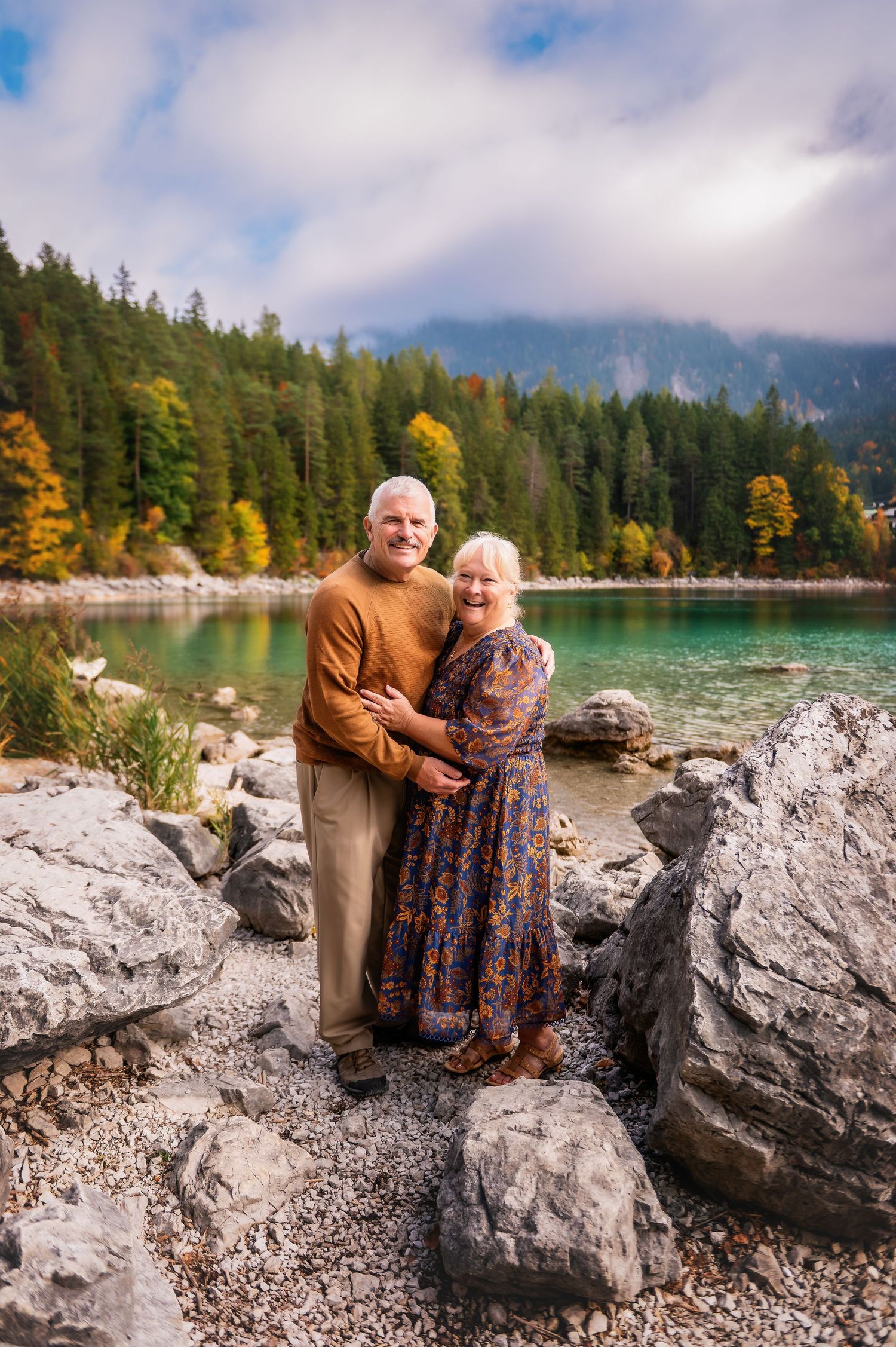 Elderly couple embracing on rocky lakeshore with turquoise water, autumn forest, and misty mountains in background.
