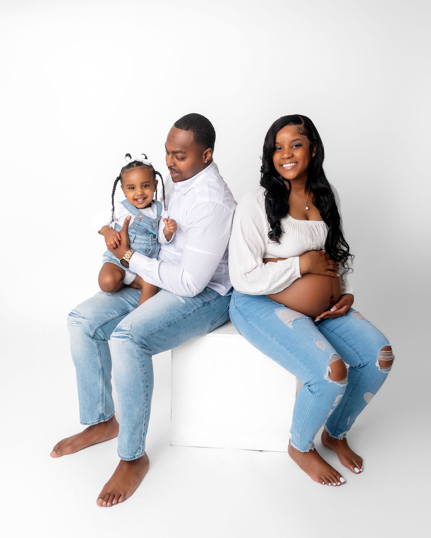 Family maternity photo with couple and toddler in casual denim against white background.