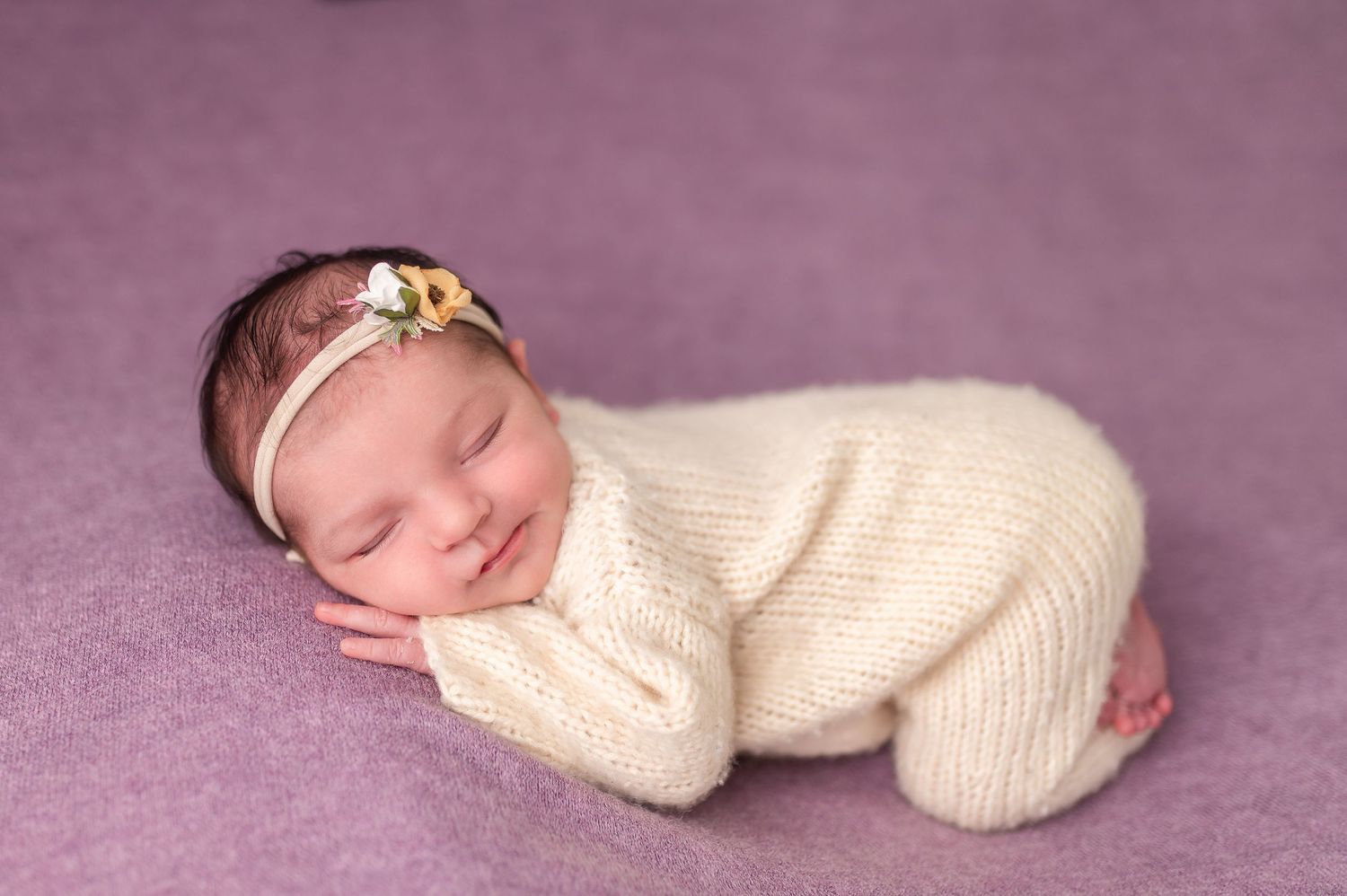 Newborn baby in cream knit outfit sleeping on purple backdrop.