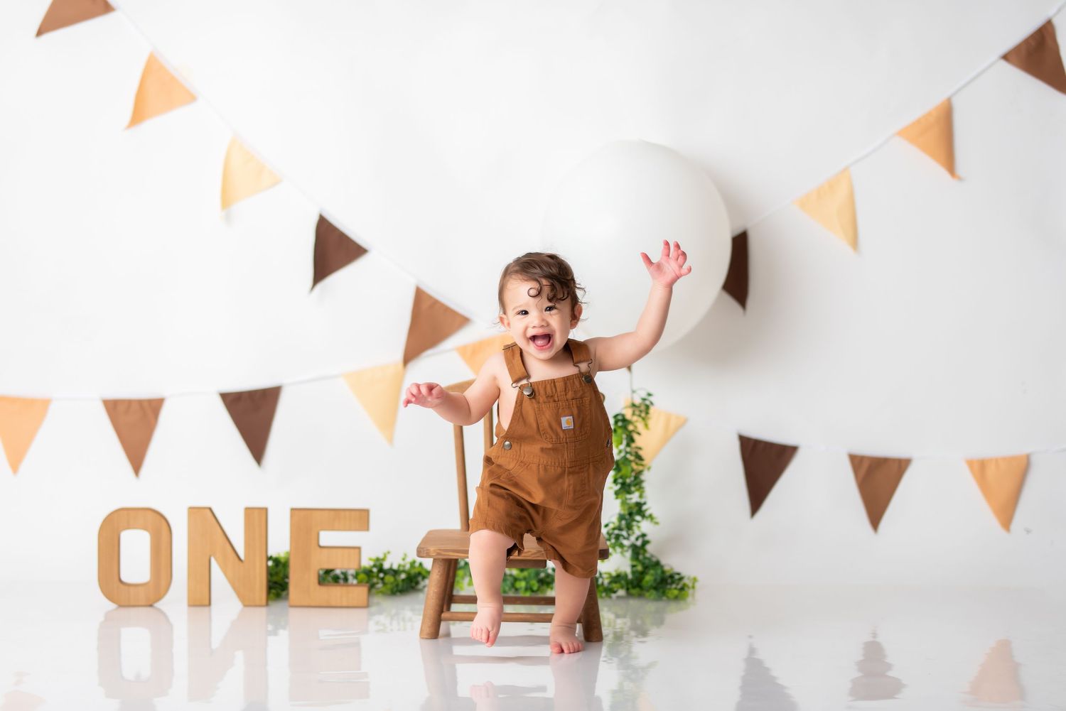 First birthday photo setup with brown overalls against white background with orange triangle bunting.