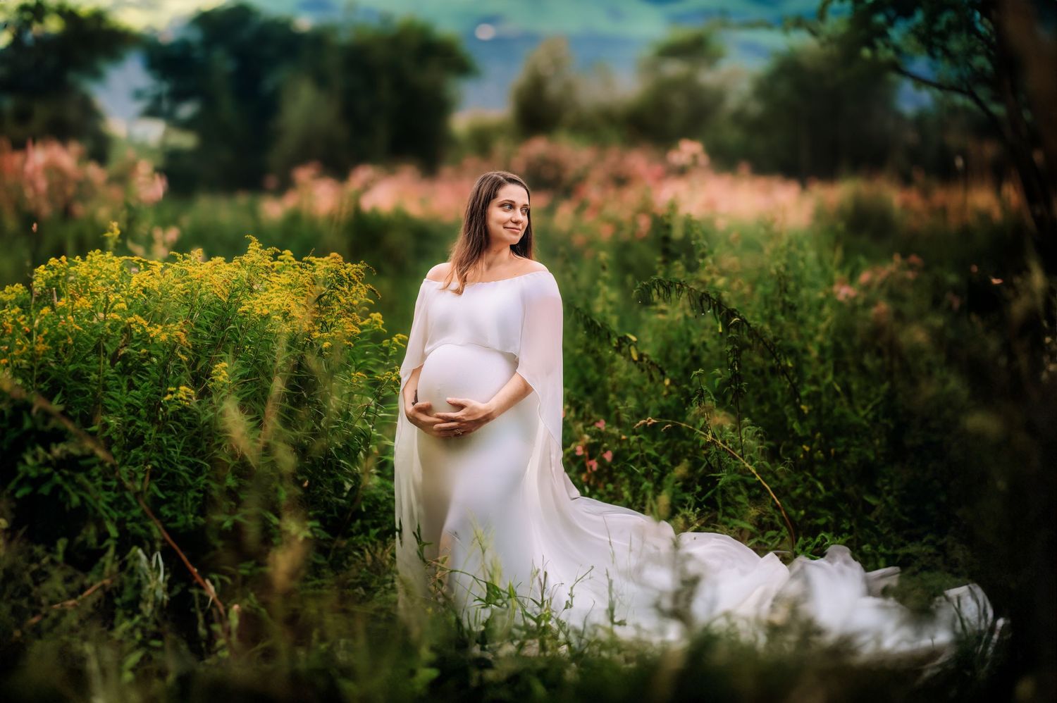Ethereal maternity portrait in white dress among wildflowers at golden hour.