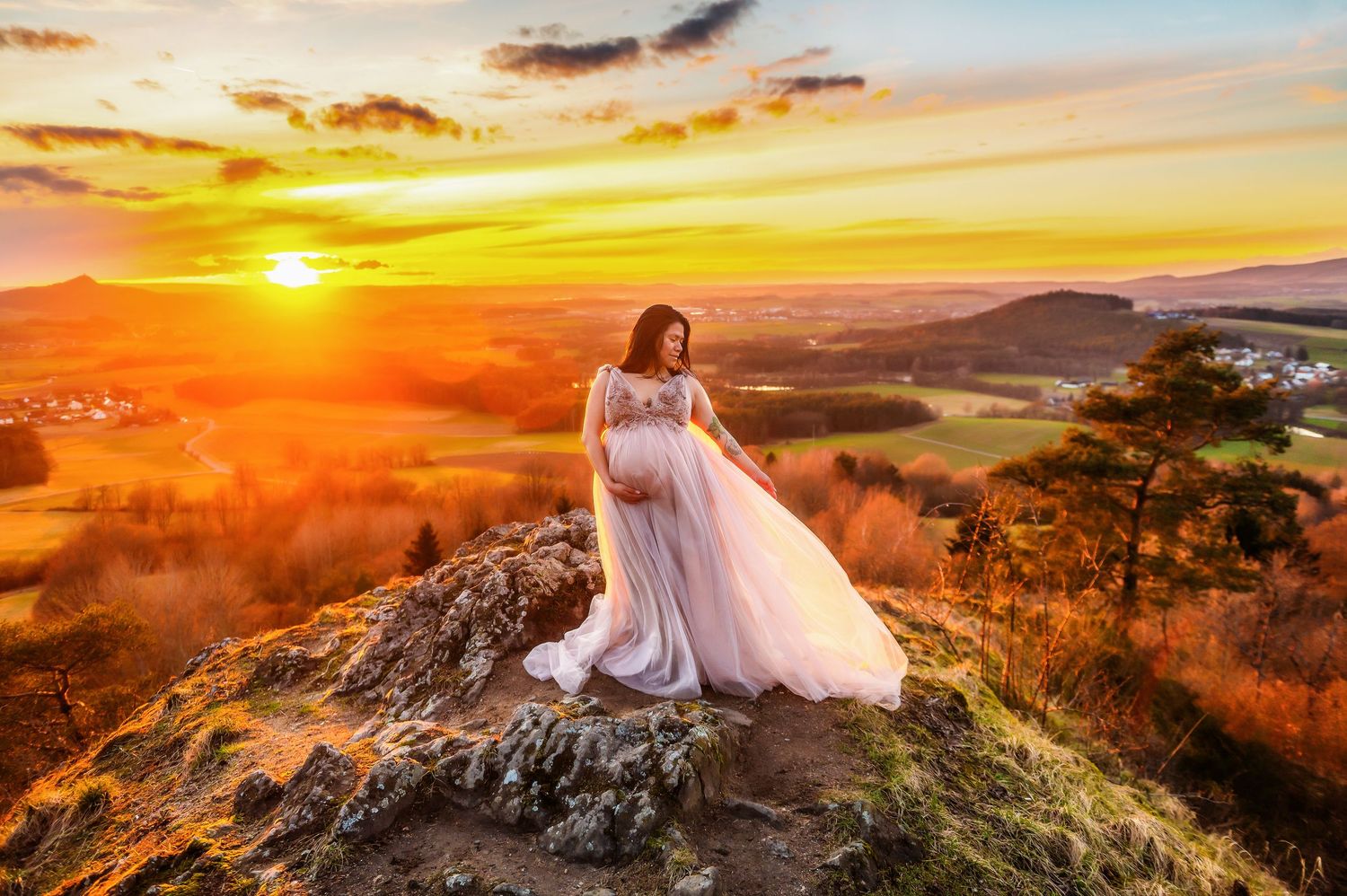 Dramatic maternity photo of figure in flowing white gown standing on mountain peak at sunset.