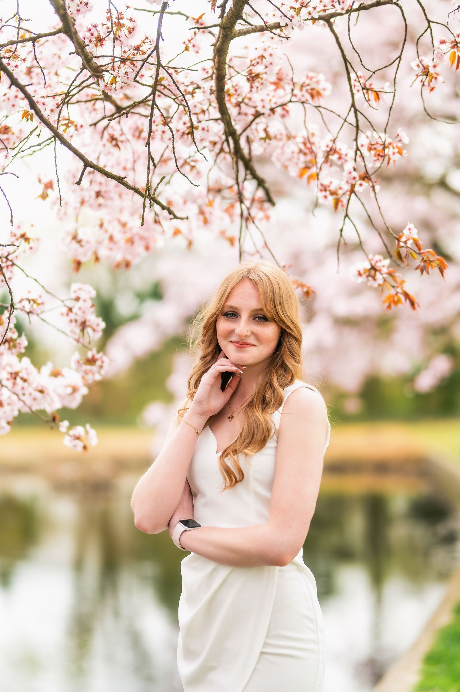 A graduate poses for portraits under blooming cherry trees beside a pond.