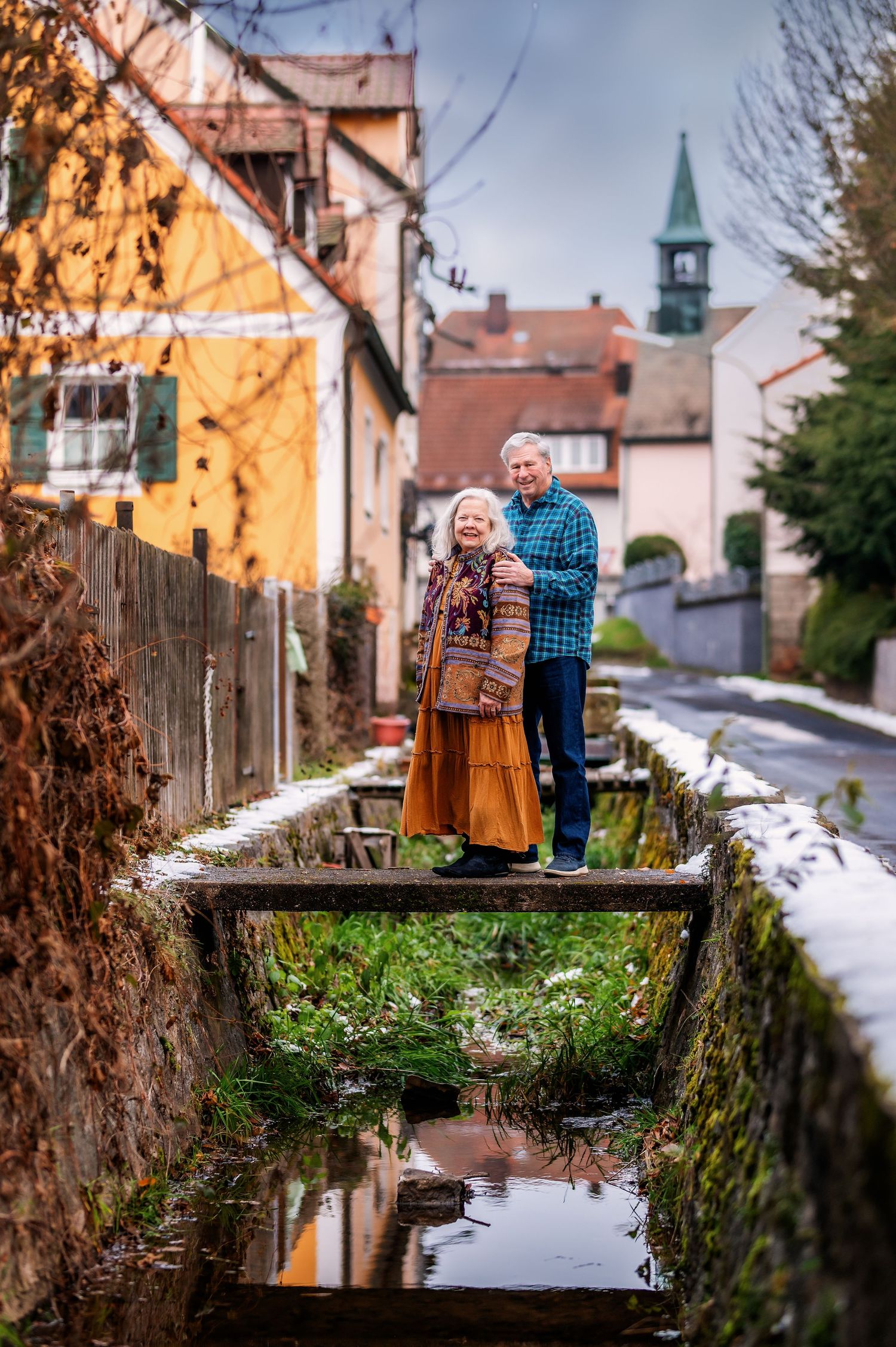Couple stands together on a rustic wooden bridge over a reflective stream in a quaint European village setting.