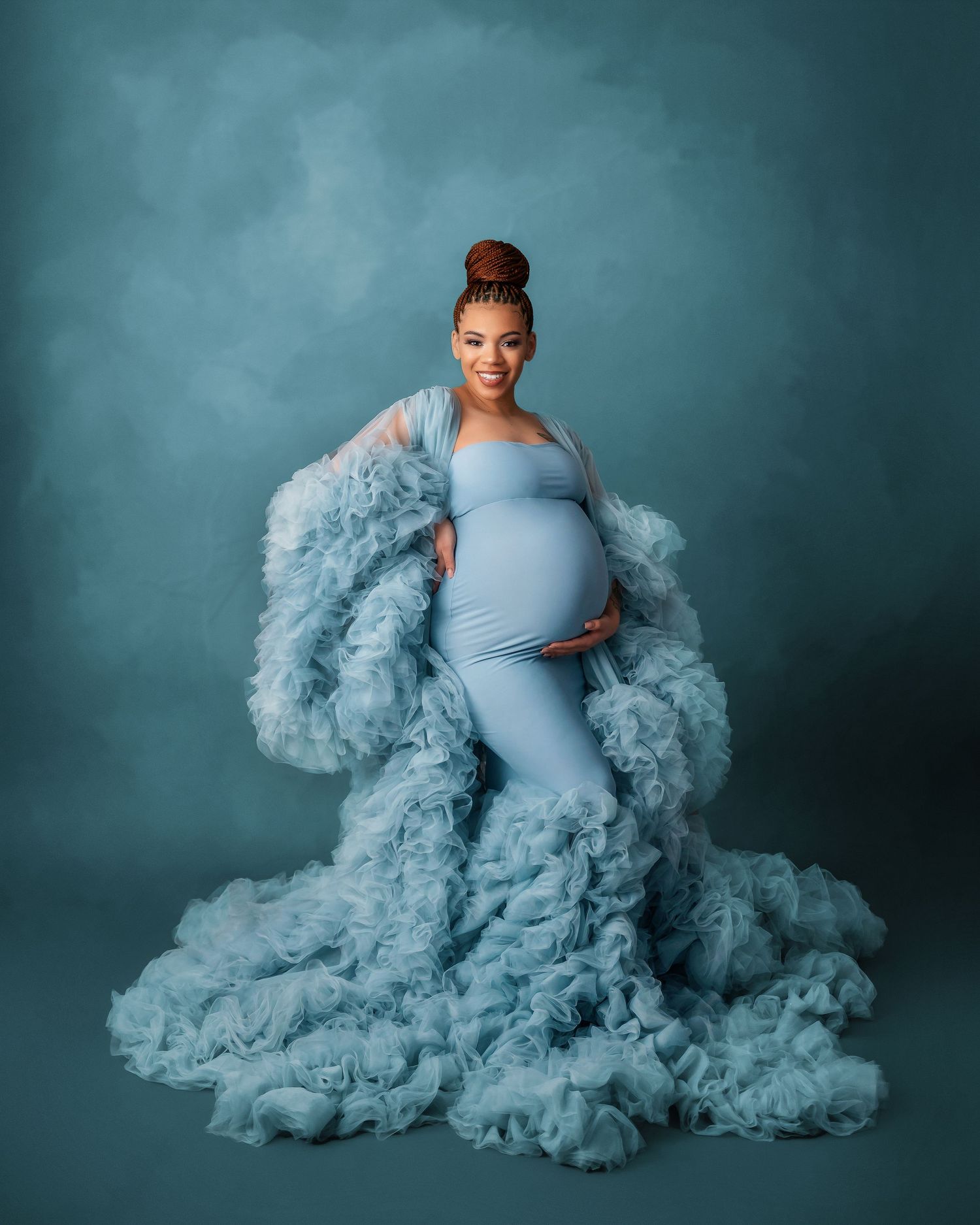 Dramatic maternity portrait with flowing blue tulle dress against teal background.