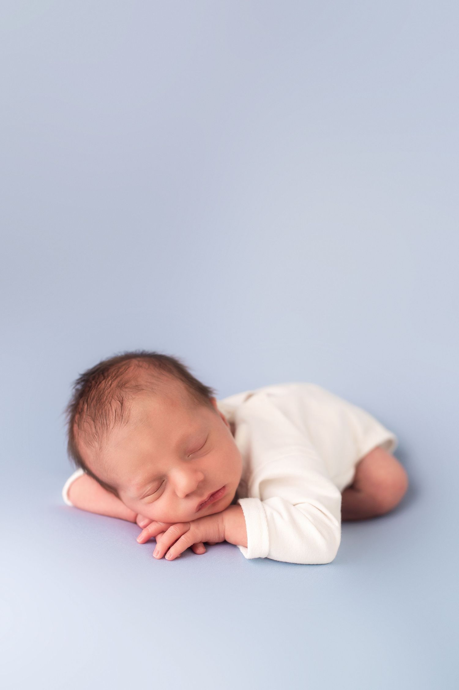 Peaceful sleeping newborn in white onesie on light blue backdrop.