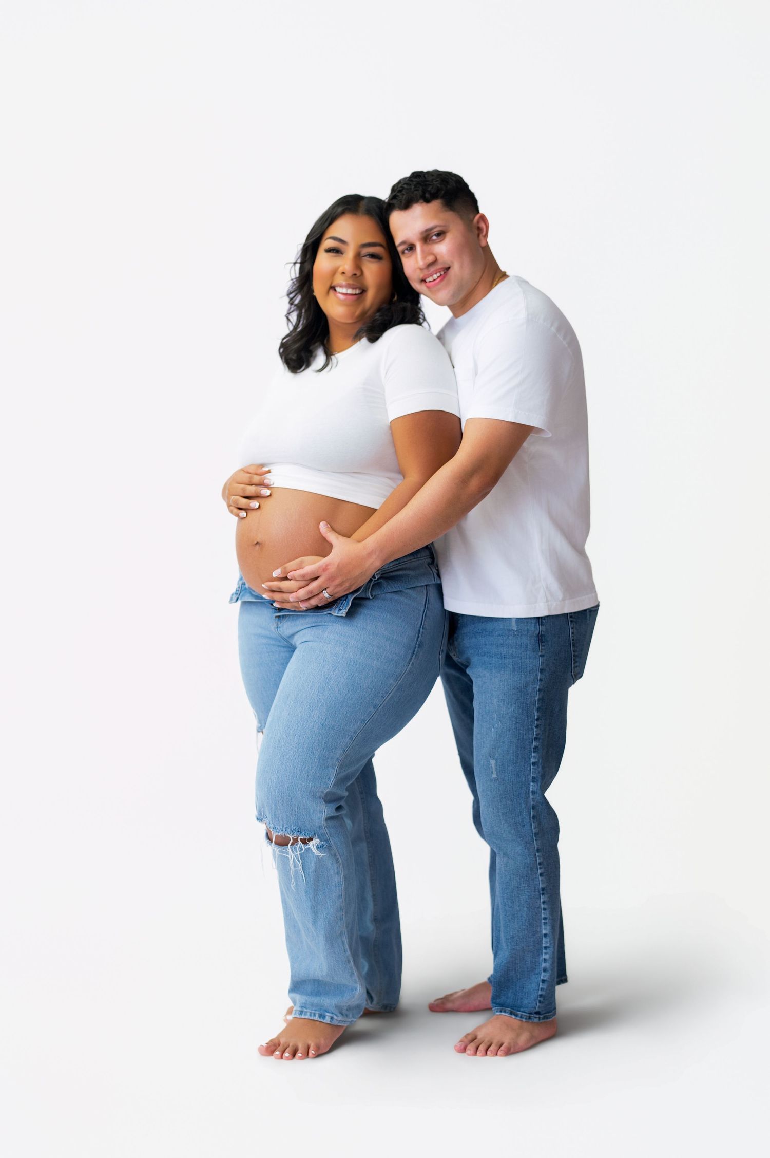 Maternity photo series showing couple in casual white and denim outfits posing together against white backdrop.