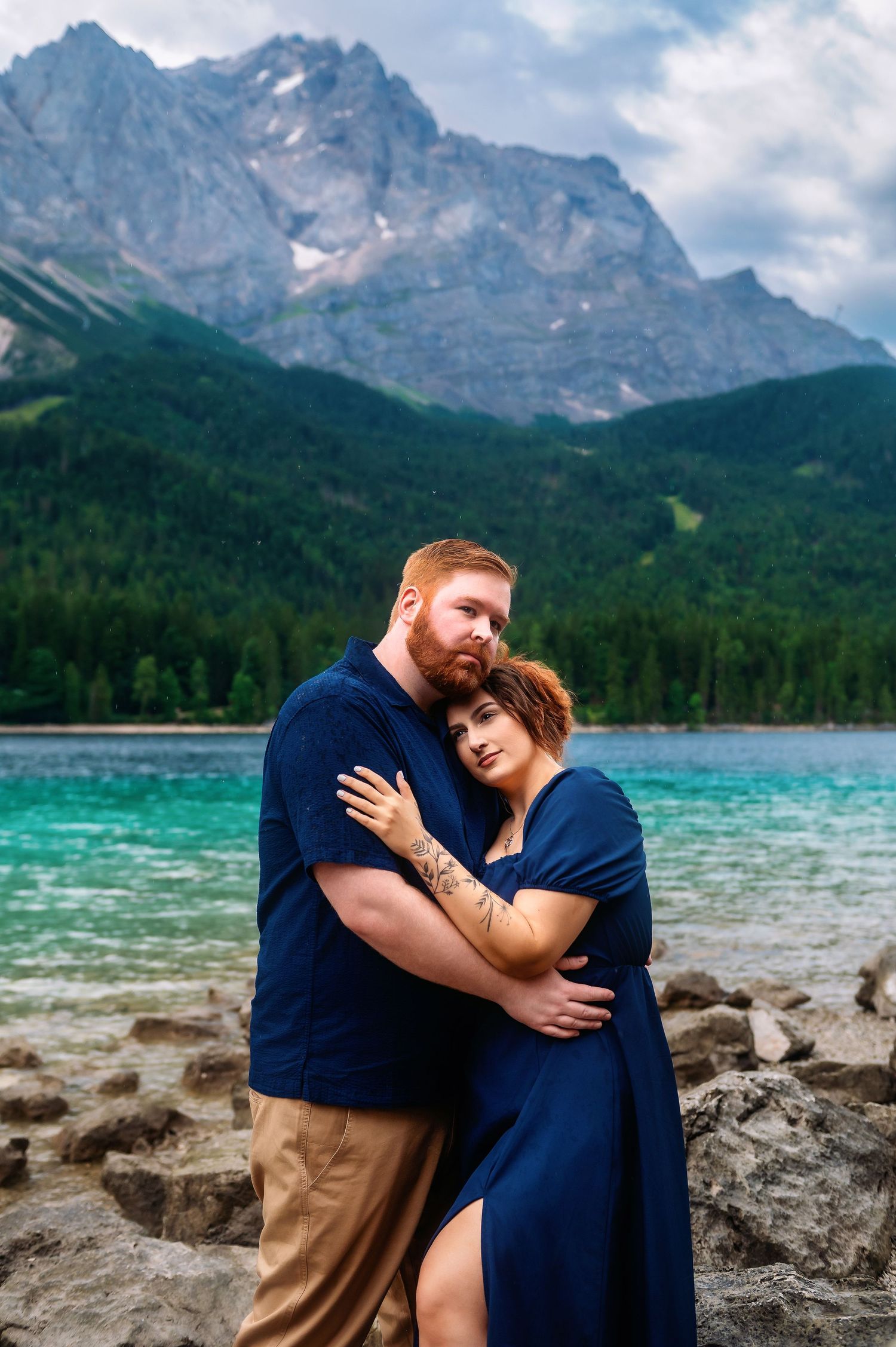 Couple in matching navy outfits embracing by a turquoise alpine lake with dramatic mountain peaks and forest backdrop.
