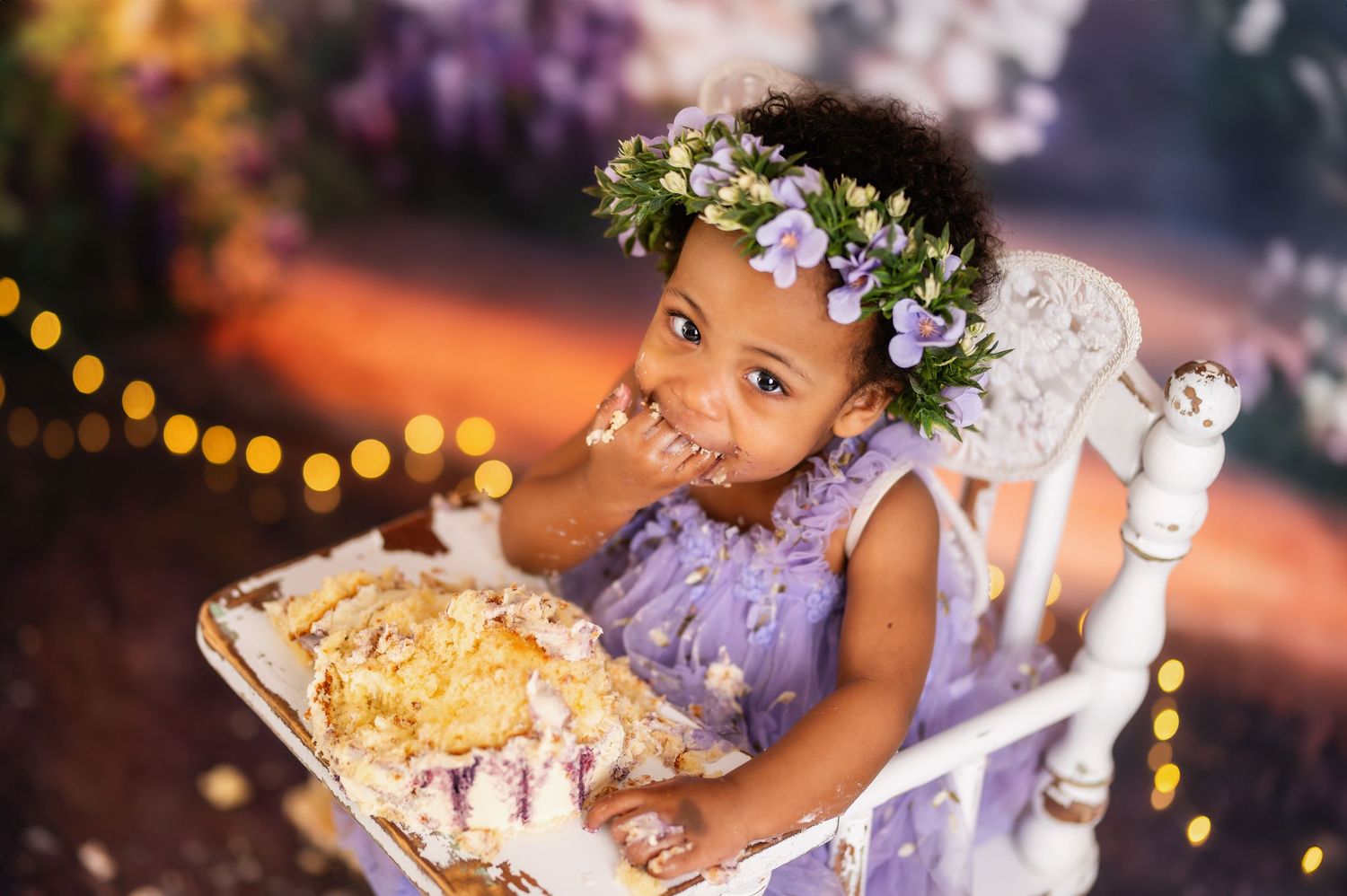 Toddler girl in purple dress and floral crown smiling during cake smash photo session outdoors.