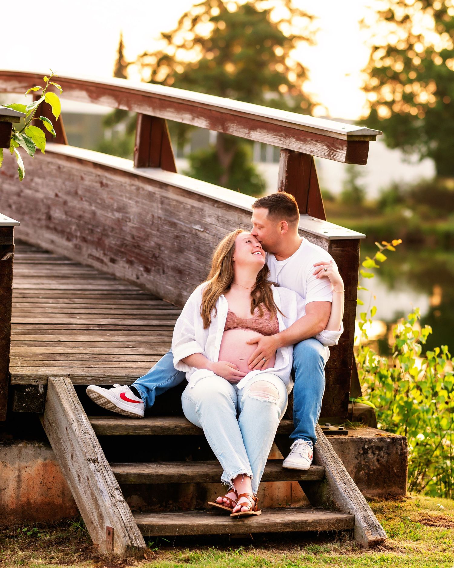 Pregnant couple sitting on wooden bridge steps at golden hour, sharing a tender moment outdoors.