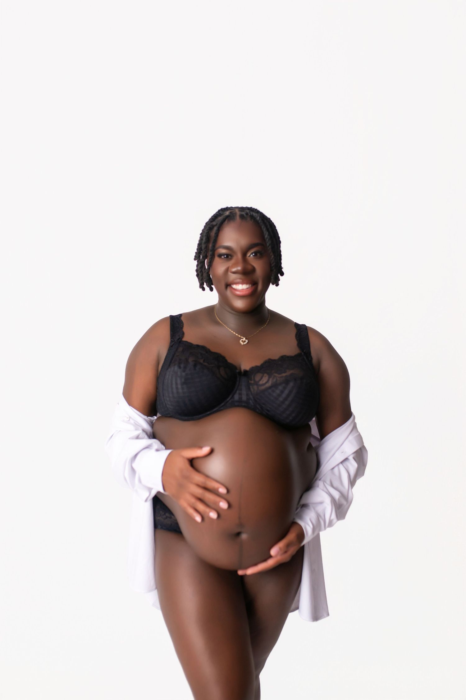 A pregnant woman in black lingerie and white shirt poses confidently against a white background.