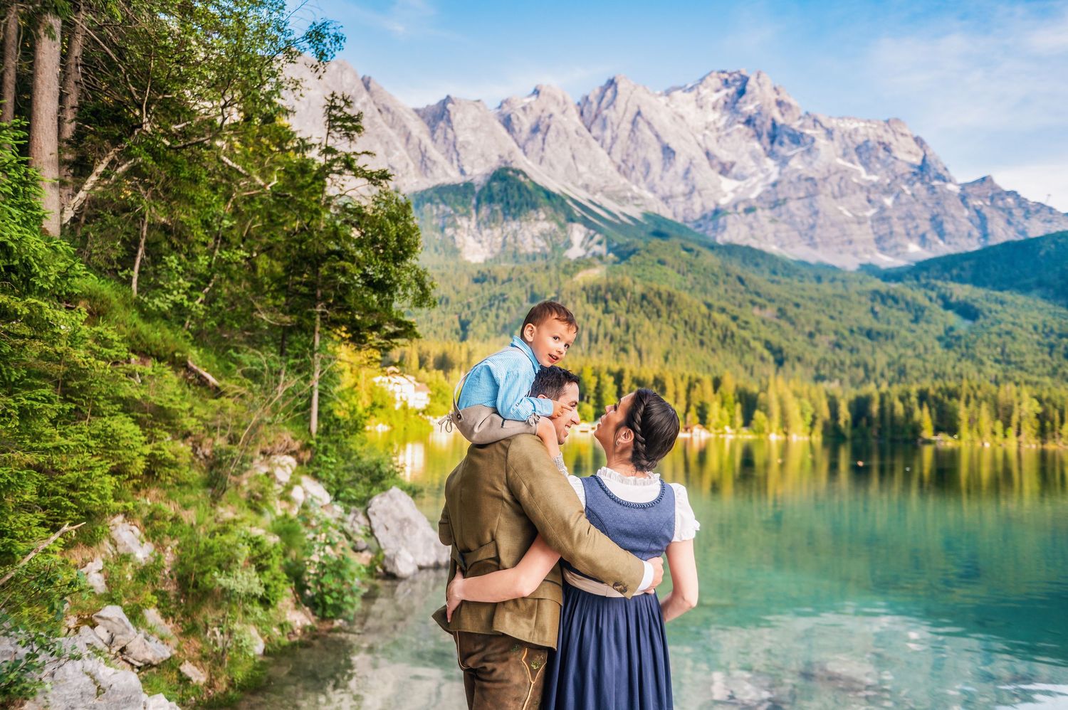 A couple stands by a tranquil mountain lake surrounded by majestic peaks and evergreen forest in a scenic landscape.