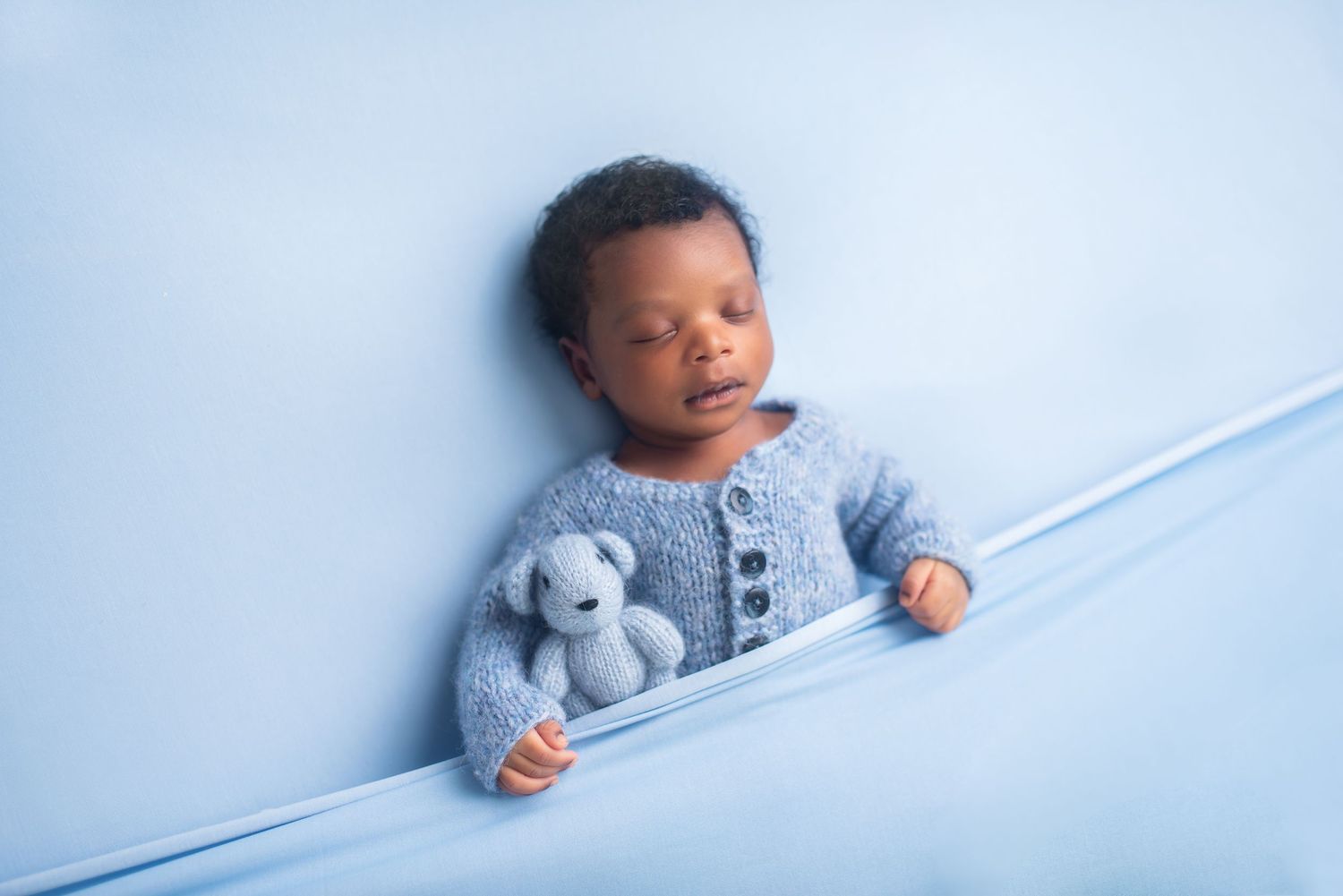 Peaceful newborn holding stuffed bear in blue knit outfit against light blue backdrop.