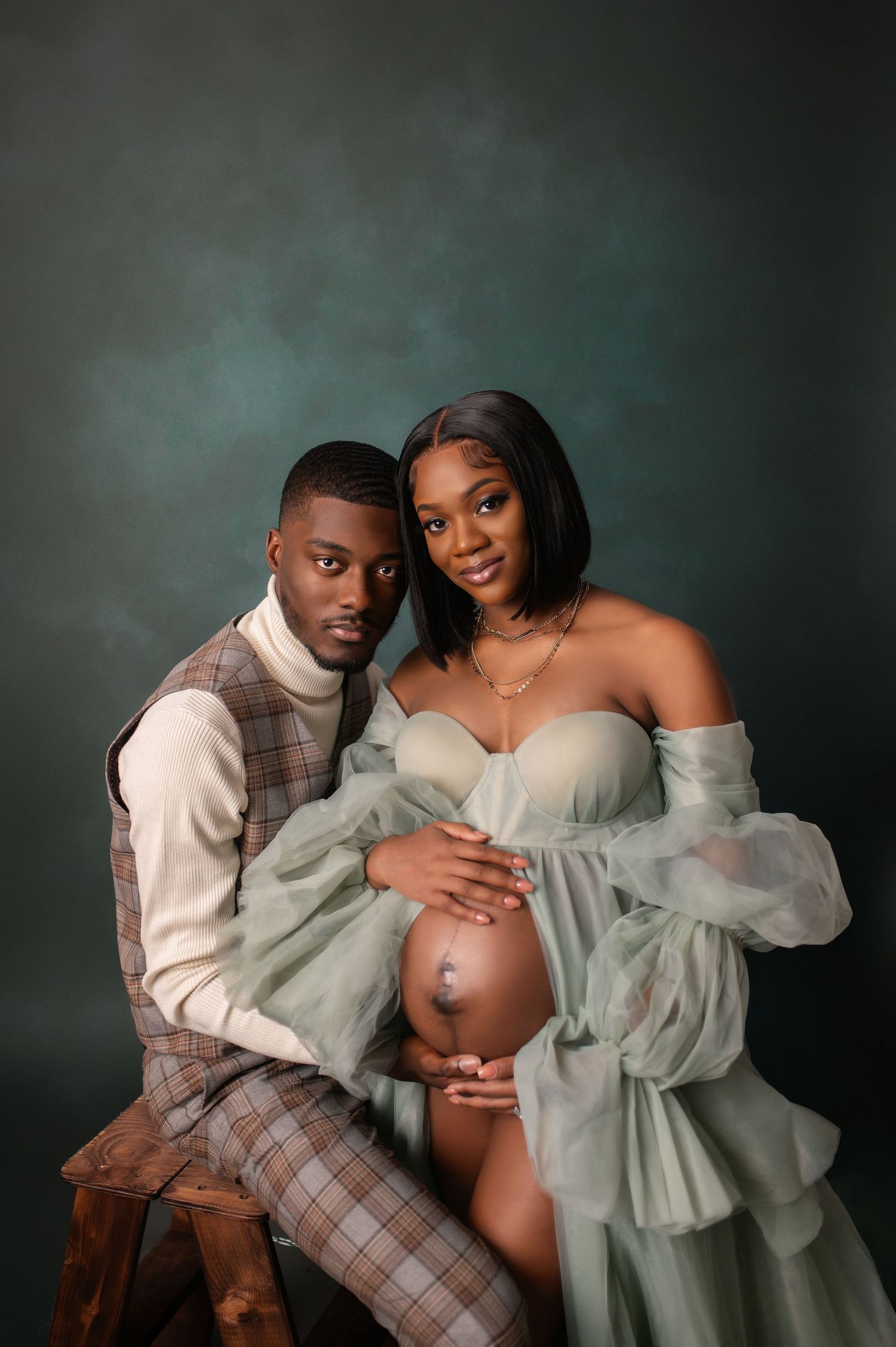 Intimate maternity portrait with couple in formal wear against dark teal backdrop.