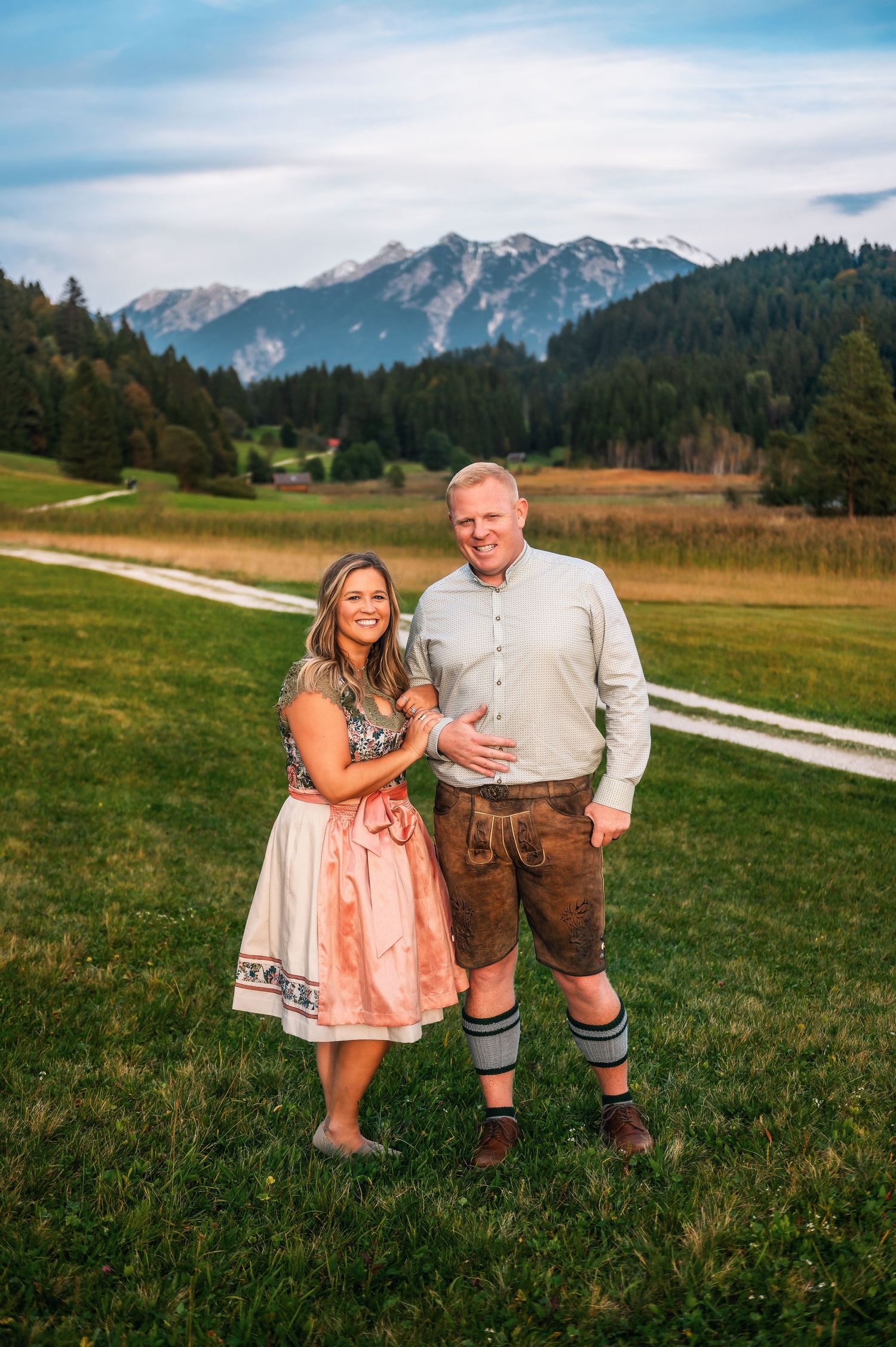Couple in traditional Bavarian dirndl and lederhosen pose together in Alpine meadow with mountains in background.