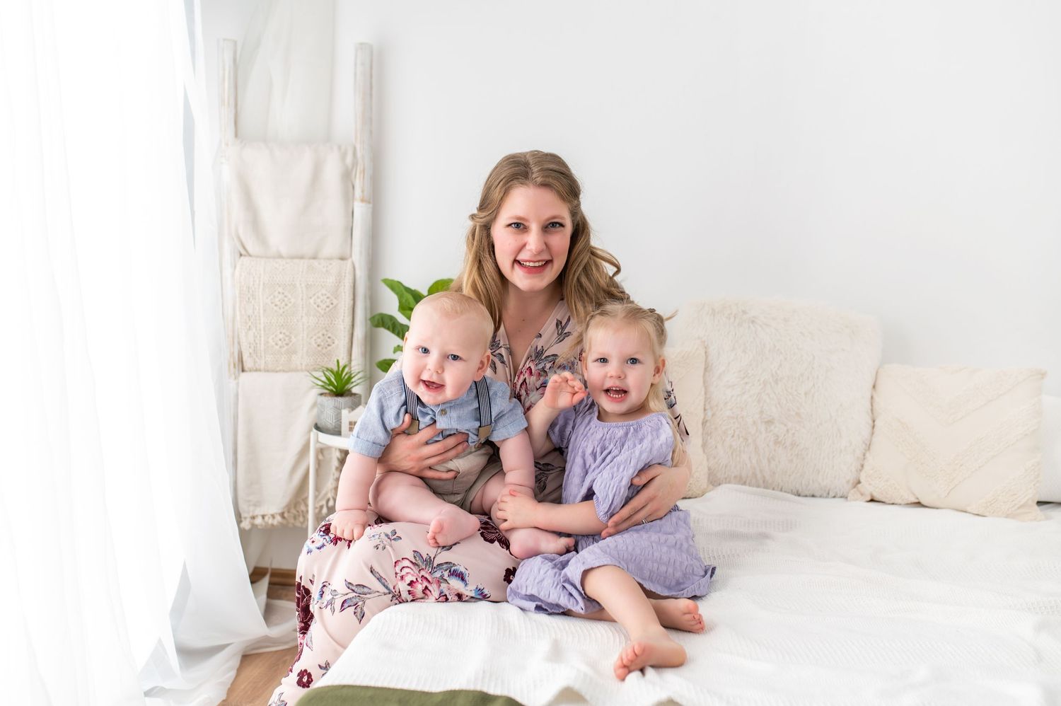 Family lying together on white bed with natural light streaming through window.