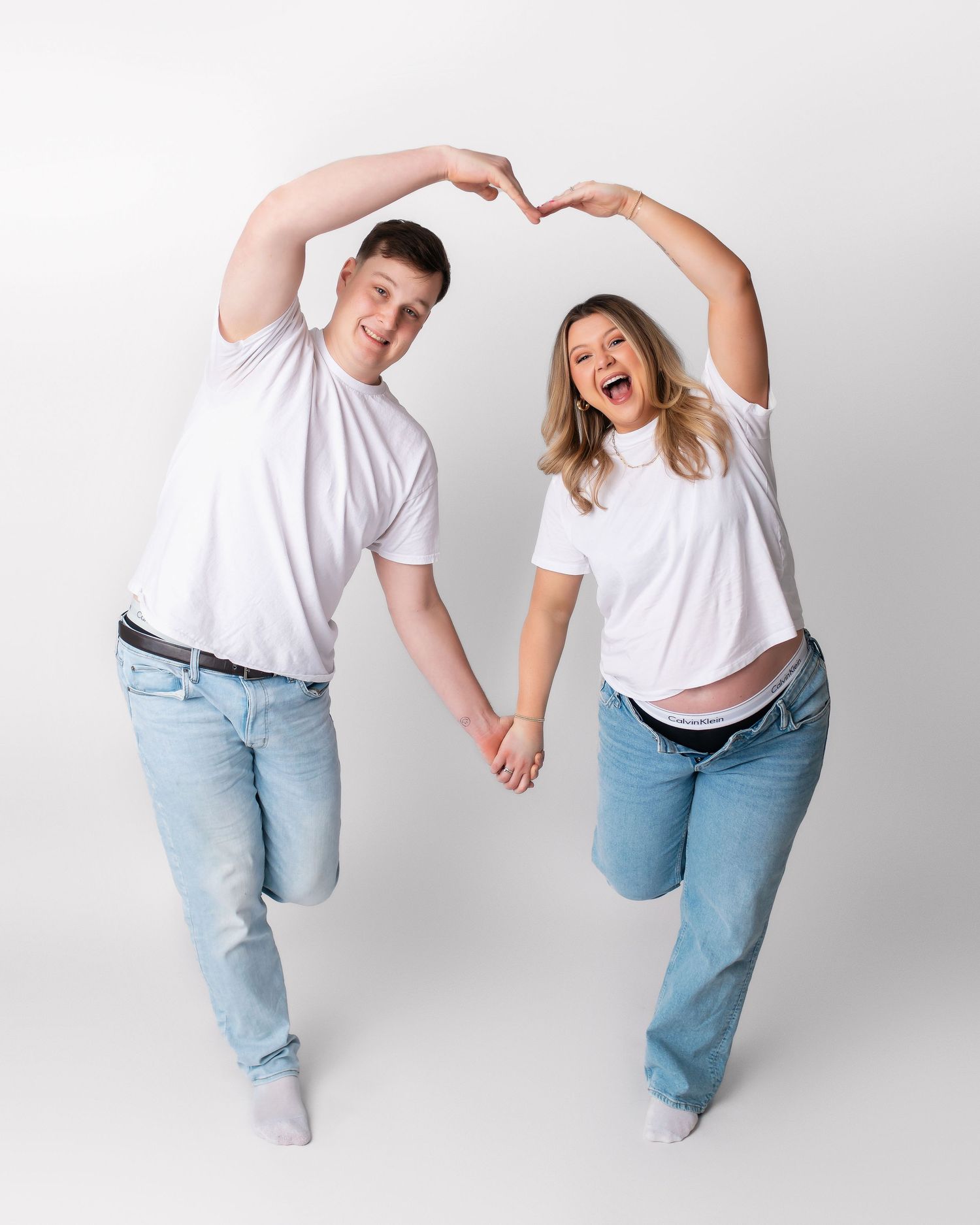 Couple in white shirts and jeans make heart shape with arms in playful studio pose.