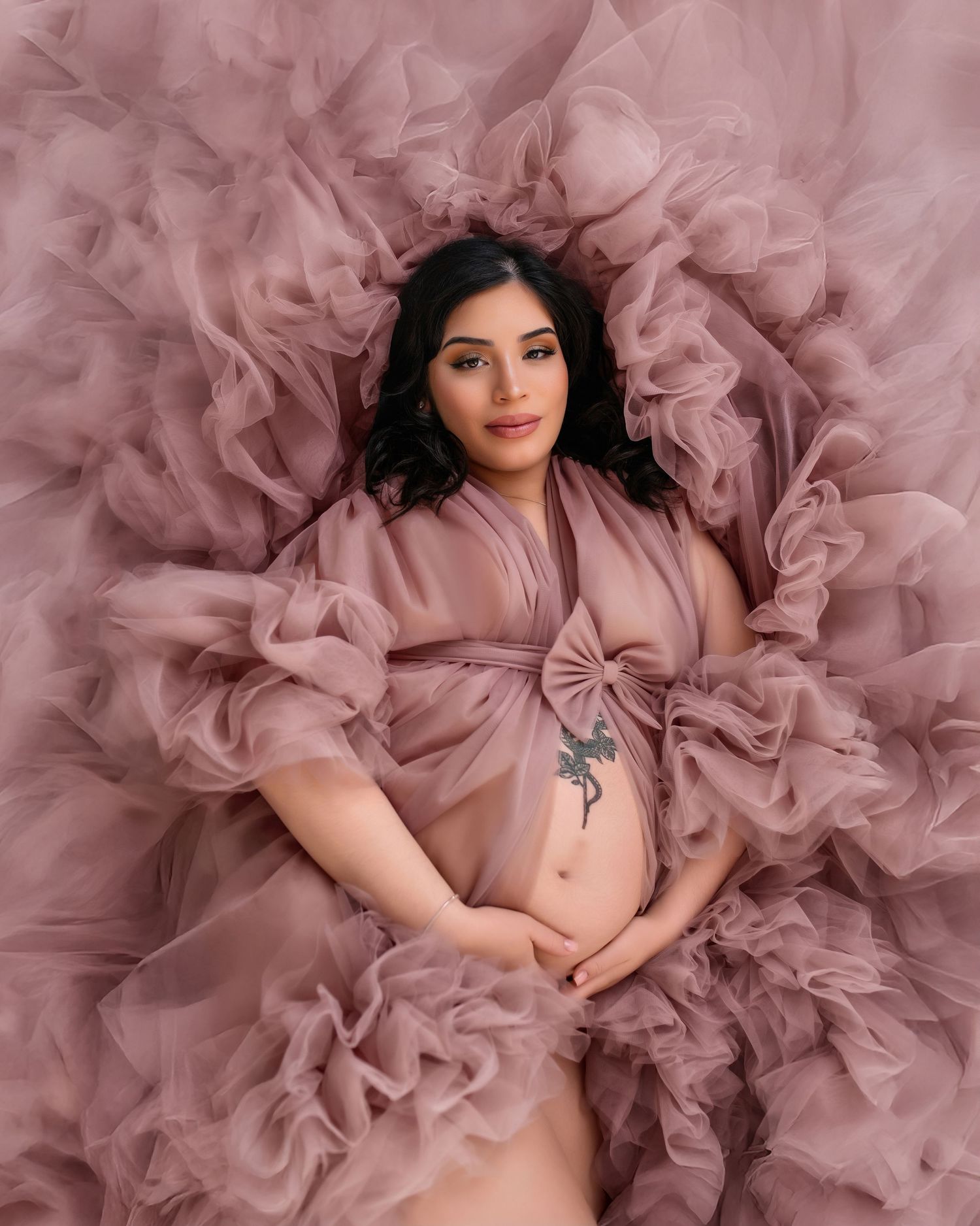 Pregnant woman in dusty rose tulle robe lying gracefully, cradling baby bump in elegant maternity photo.