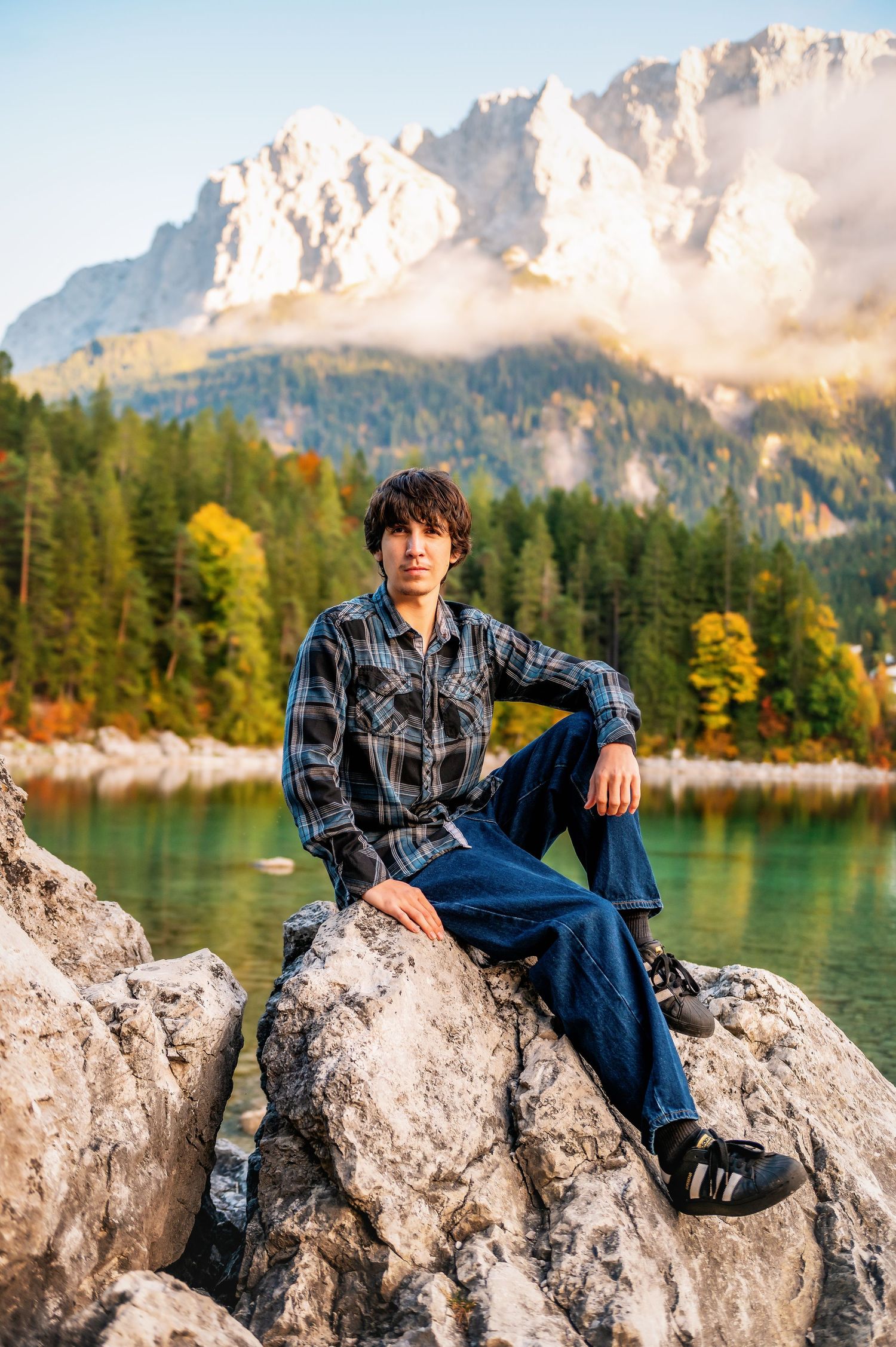 Scenic mountain landscape with lake and autumn trees reflected in calm waters as someone relaxes on rocky shore.