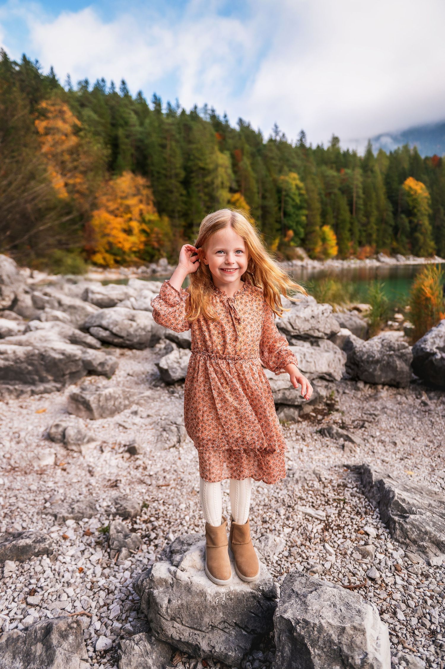 A child in a floral dress plays on rocky terrain near evergreen trees and autumn foliage by a lake.