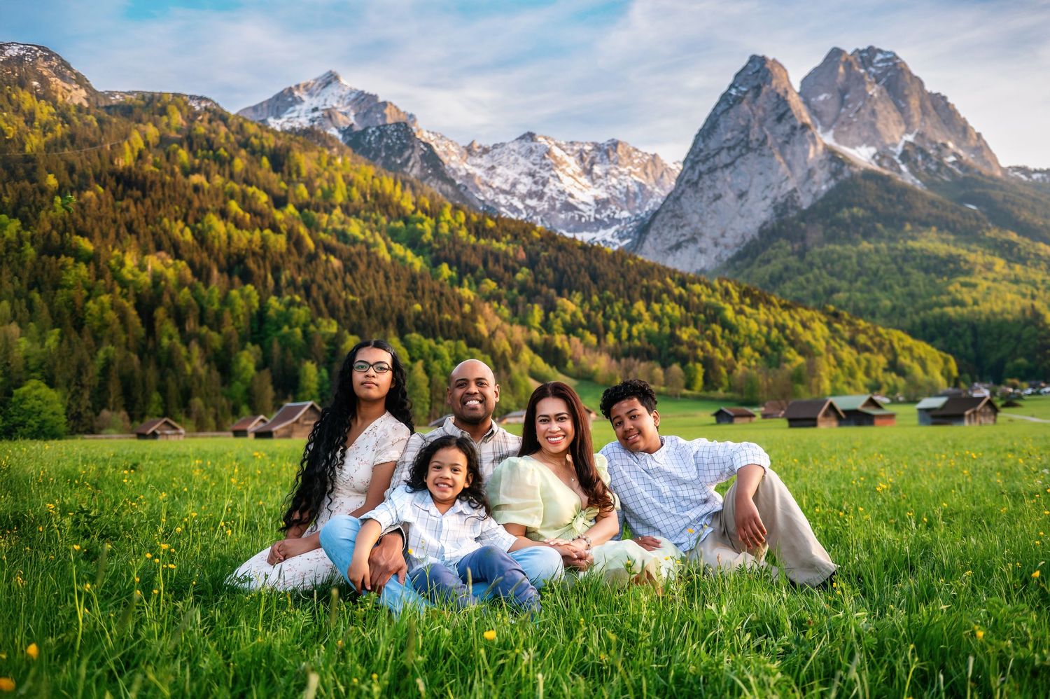 Family of five sitting on green grass with snow-capped mountains and autumn trees in the background.