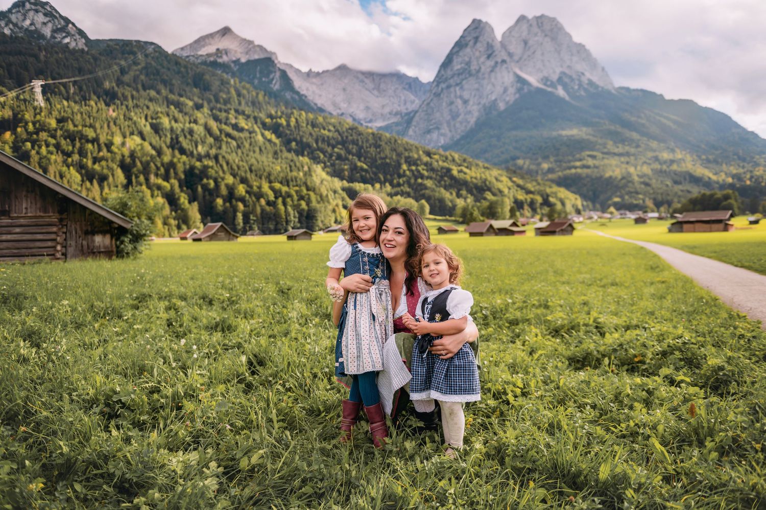 Mother and two daughters in traditional dress pose in Alpine meadow with dramatic mountain peaks and green forests backdrop.