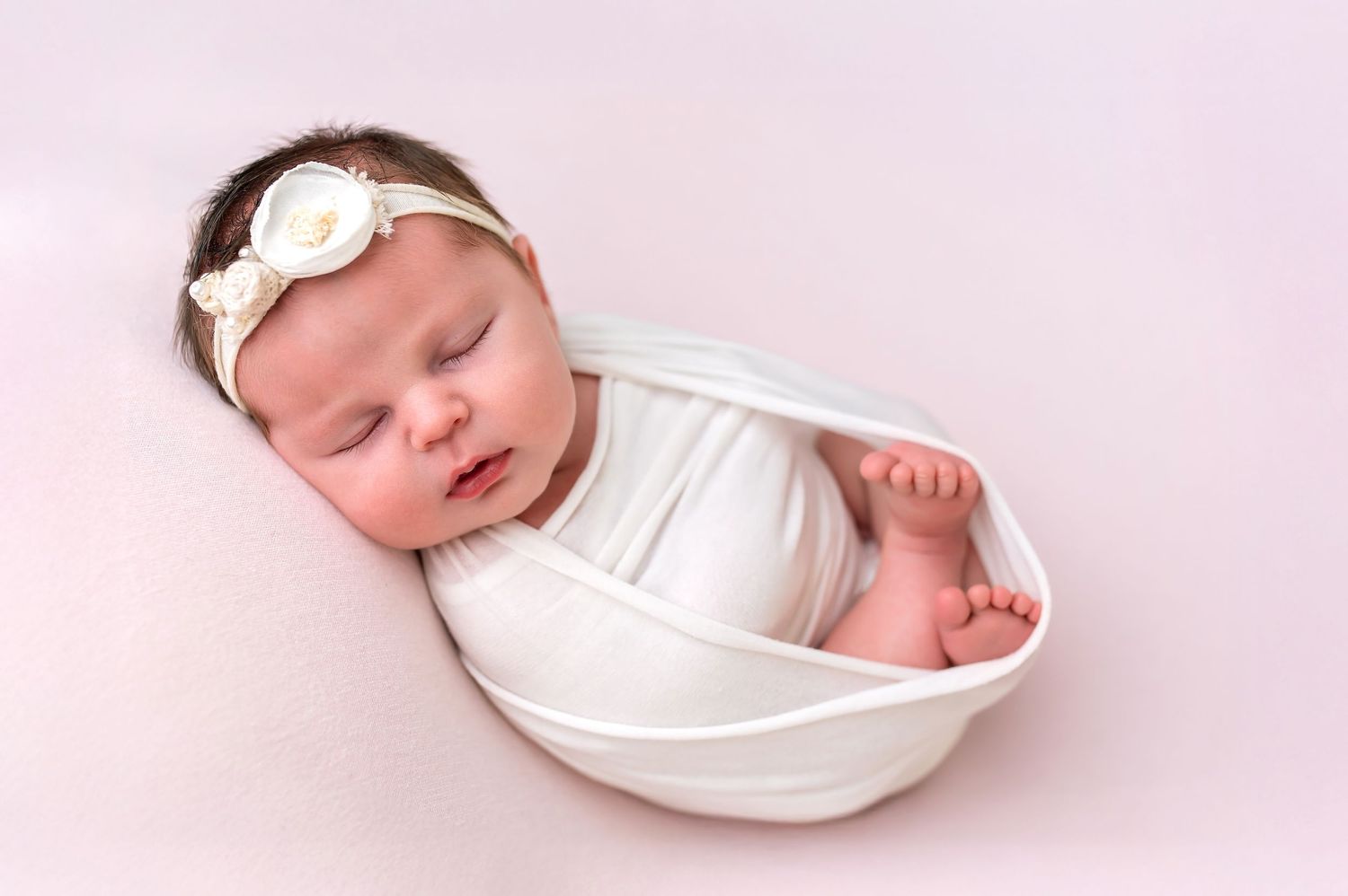 A sleeping newborn baby wearing a white bow headband rests peacefully in a curved white bowl against a pink background.