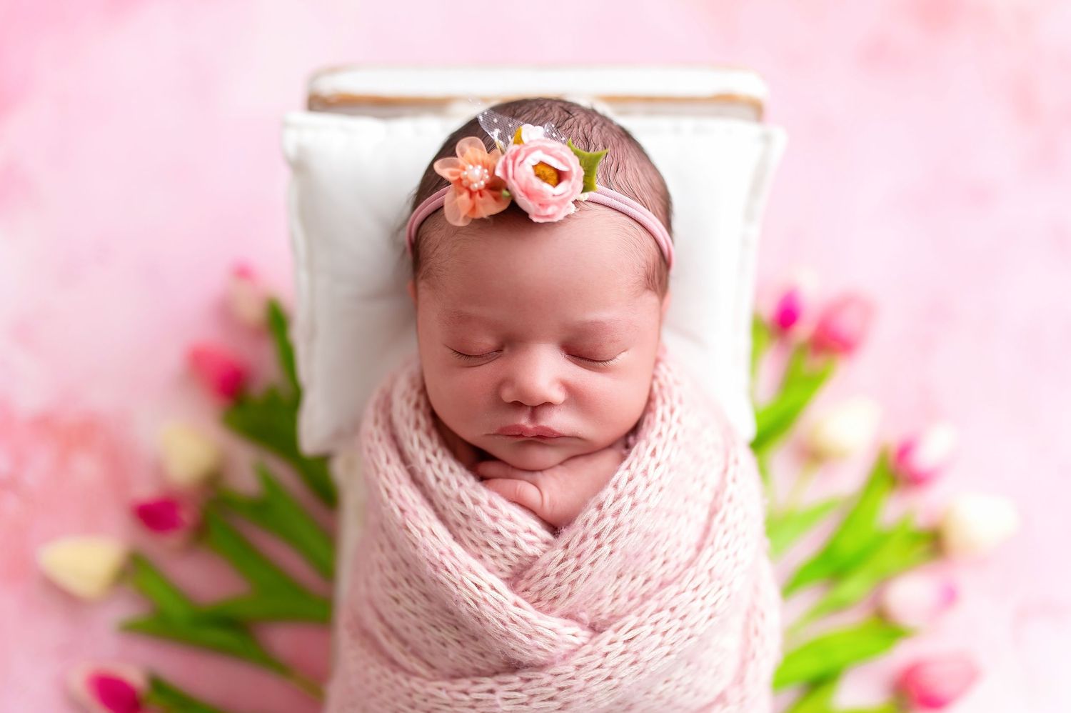 Newborn with floral headband wrapped in pink swaddle with tulips in background.