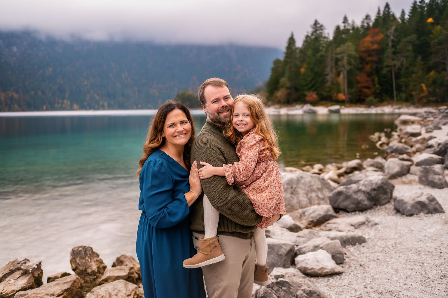 Family of three smiling by a scenic mountain lake with rocky shores and colorful autumn forest backdrop.