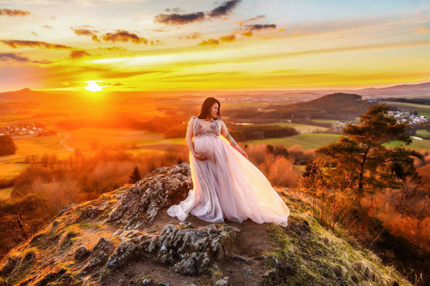Pregnant woman in flowing white gown standing on rocky hilltop at golden sunset with scenic valley landscape.