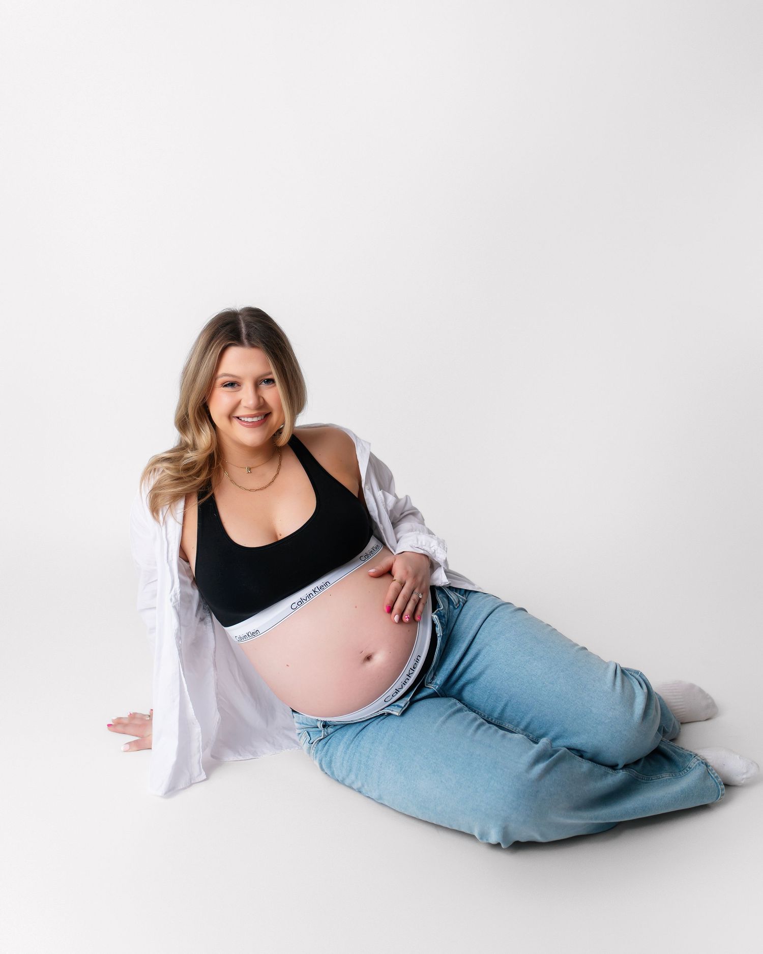 Smiling pregnant woman in black Calvin Klein bralette, open white shirt, and light blue jeans posing on white studio background.