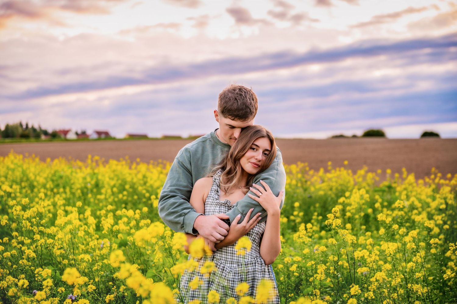 A couple embraces in a vibrant yellow canola field during golden hour with dramatic clouds in the sky.