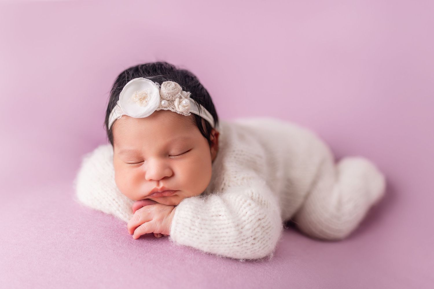 Sleeping newborn in white outfit against soft pink background with delicate styling.