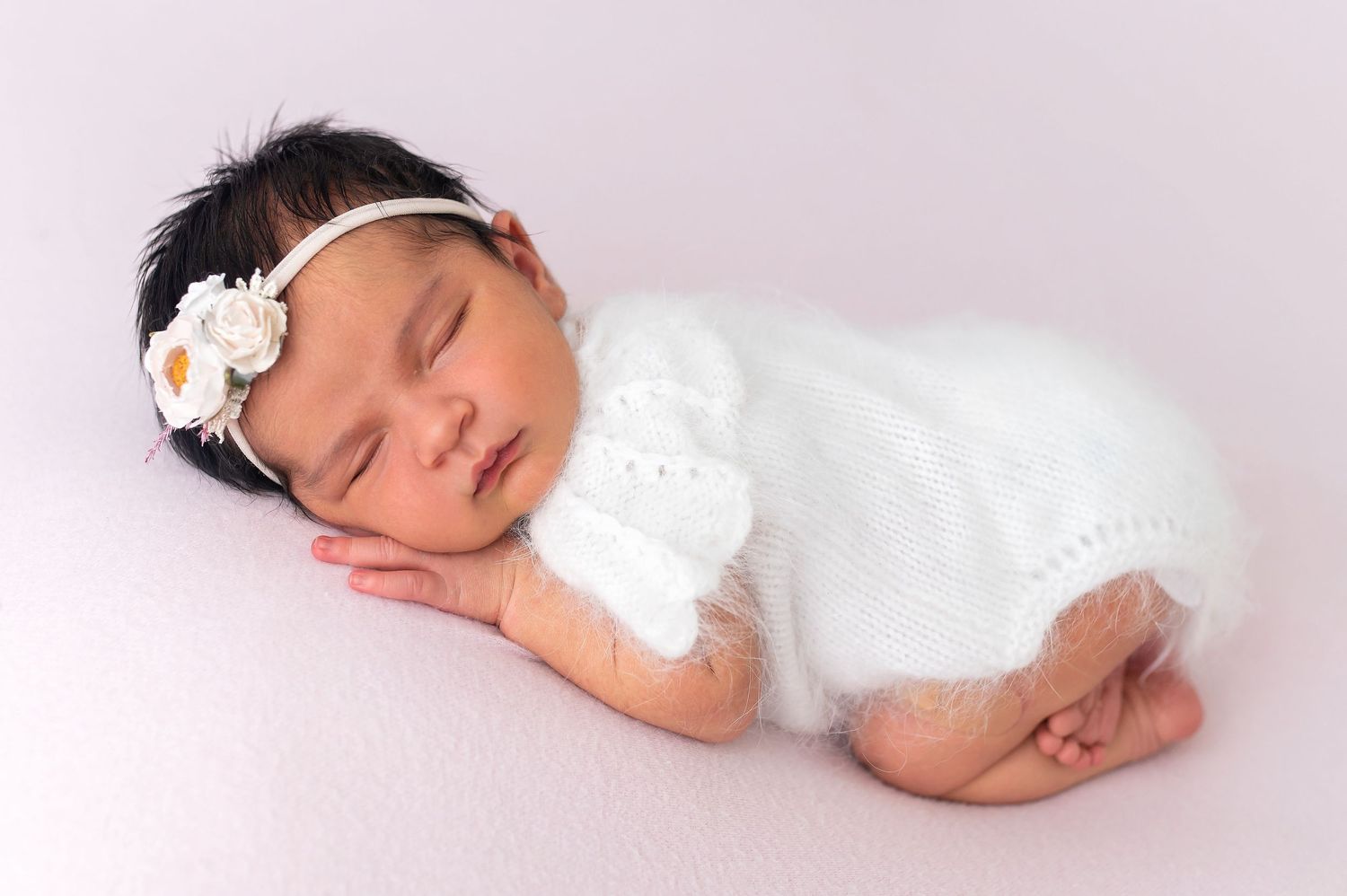 Sleeping newborn in white outfit and floral headband on pink background.