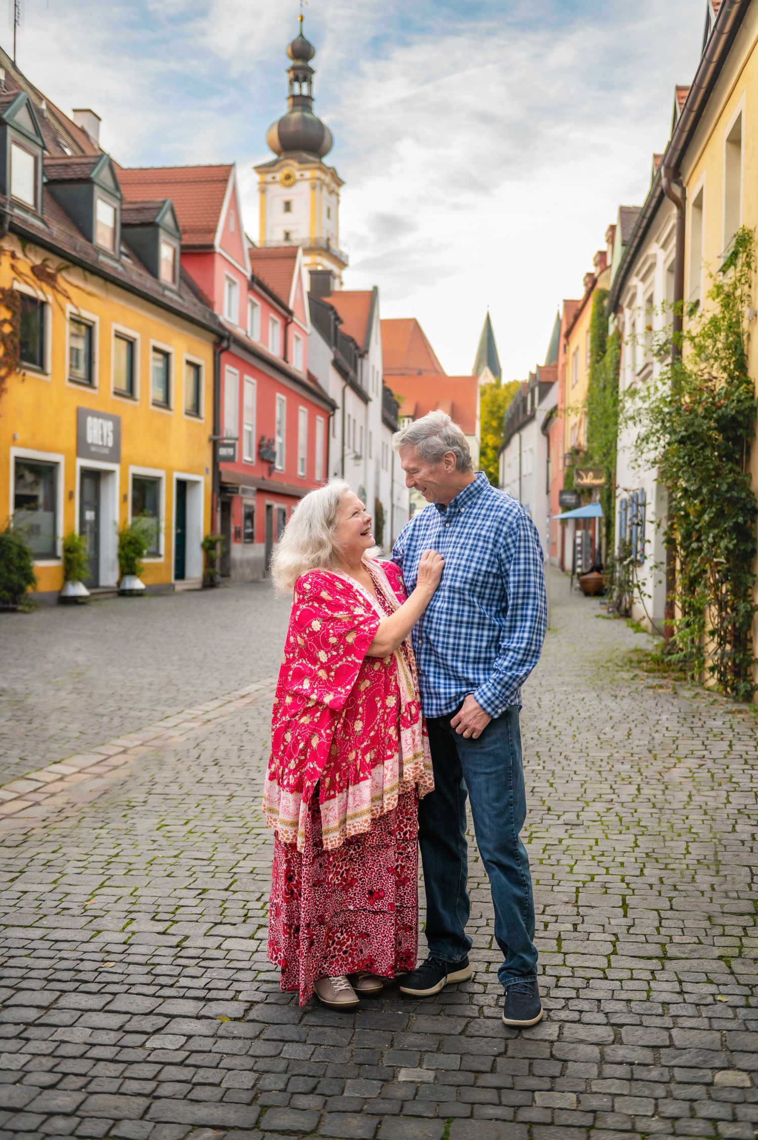 Couple embraces on cobblestone street in charming European town with colorful buildings and church spire.