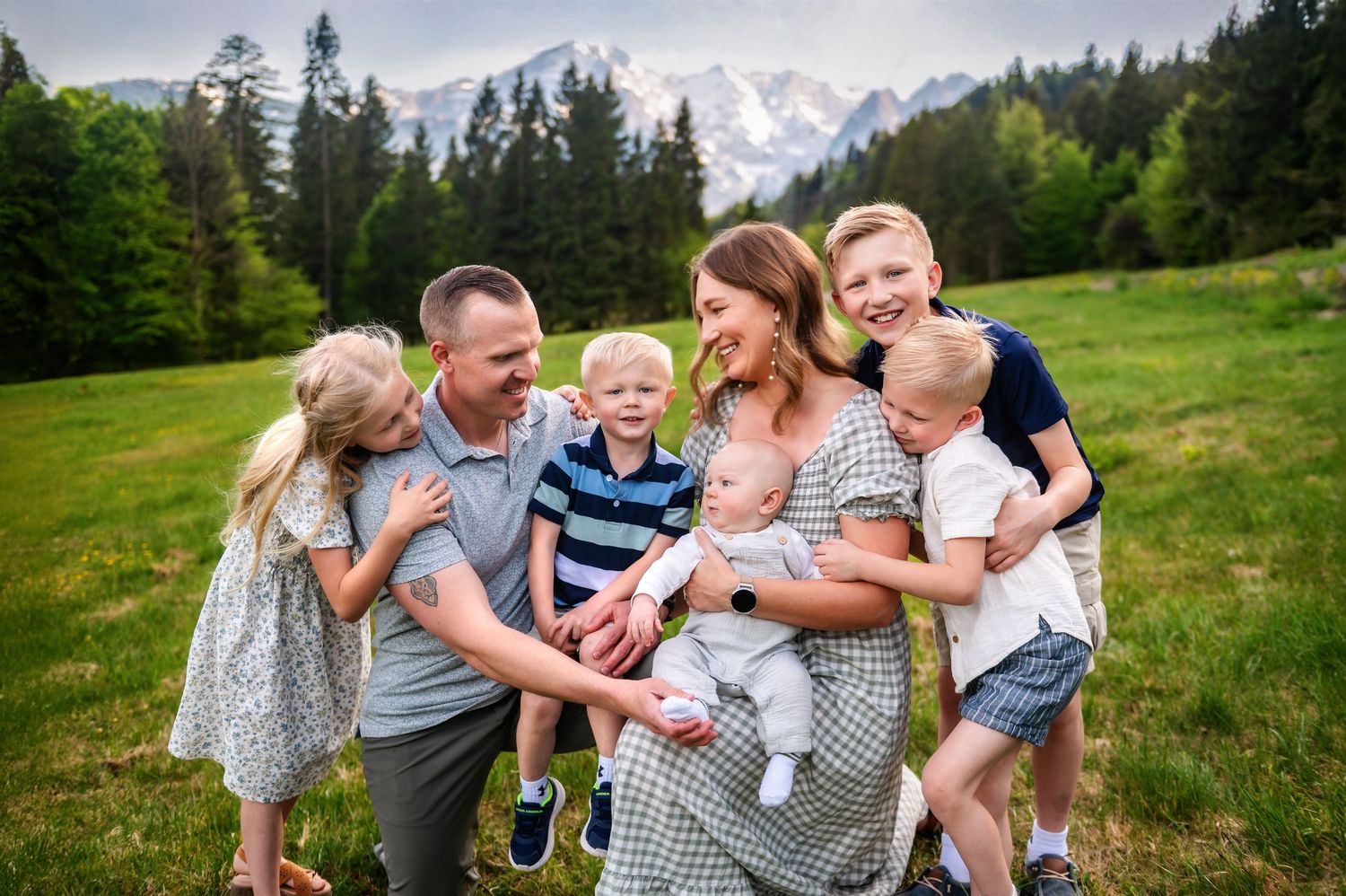 Happy family with four children posing outdoors in a green meadow with snow-capped mountains in the background.