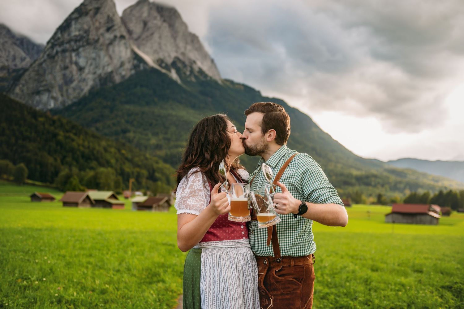 Couple in traditional Bavarian attire kissing while holding beer steins in a green alpine meadow with mountains behind.