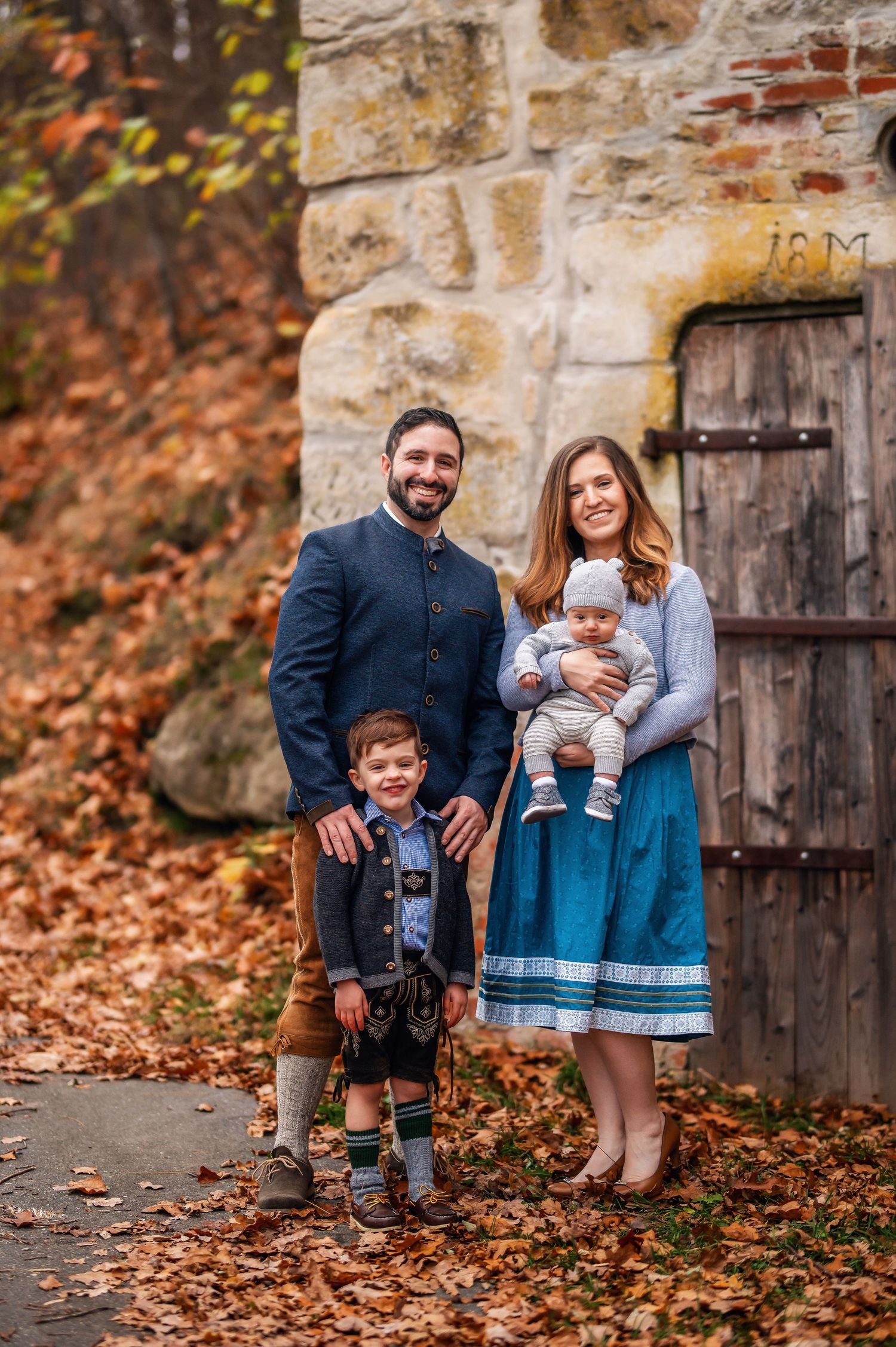 Family portrait series with traditional German outfits against stone wall with autumn leaves.