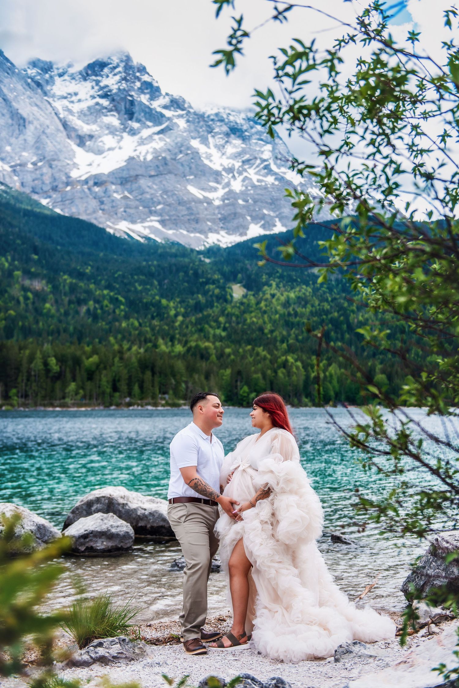 Couple in elegant attire poses by turquoise alpine lake with snow-capped mountains and forest backdrop.