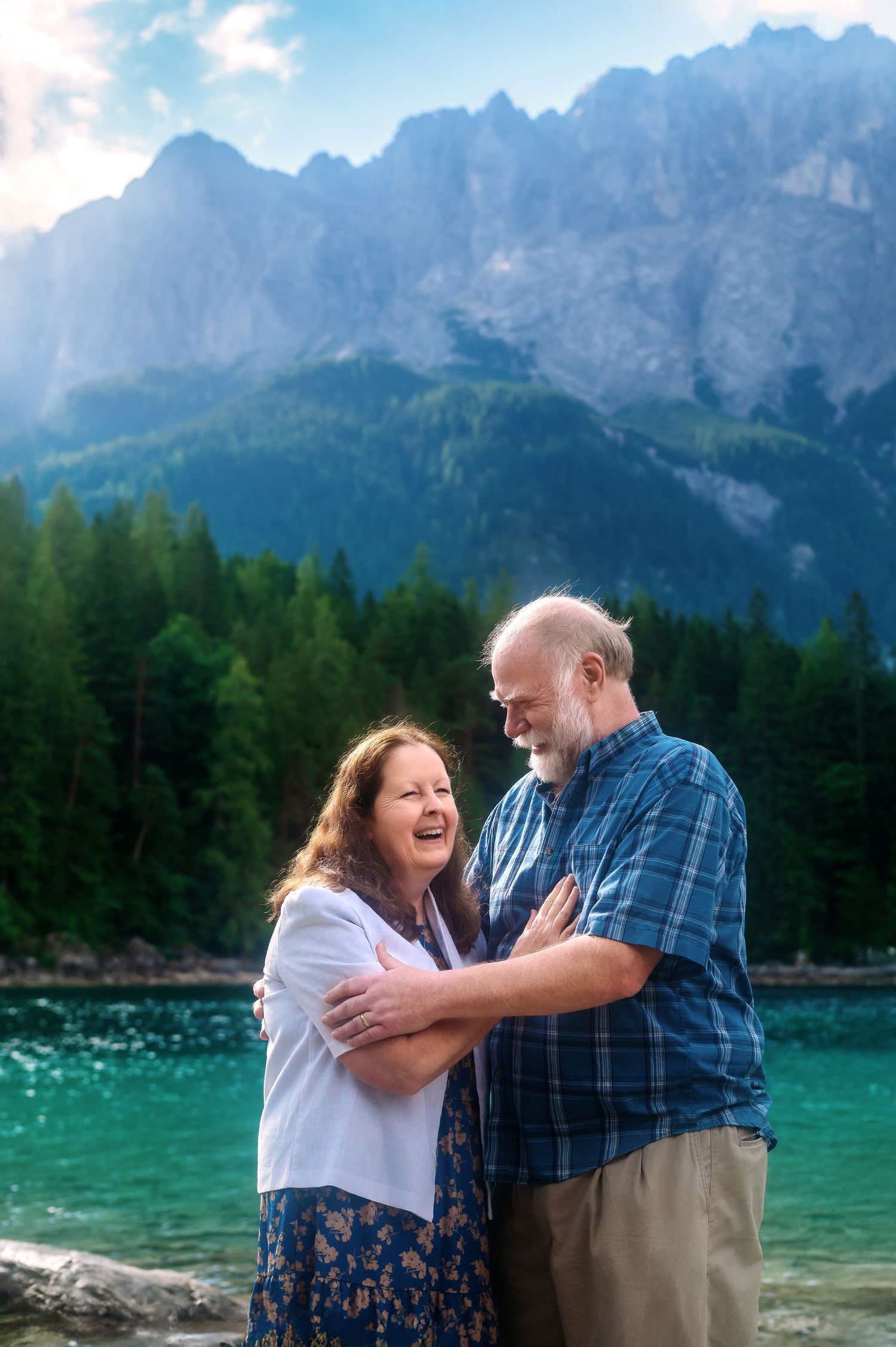 Happy senior couple embracing and laughing by a turquoise alpine lake with mountain peaks and forest backdrop.