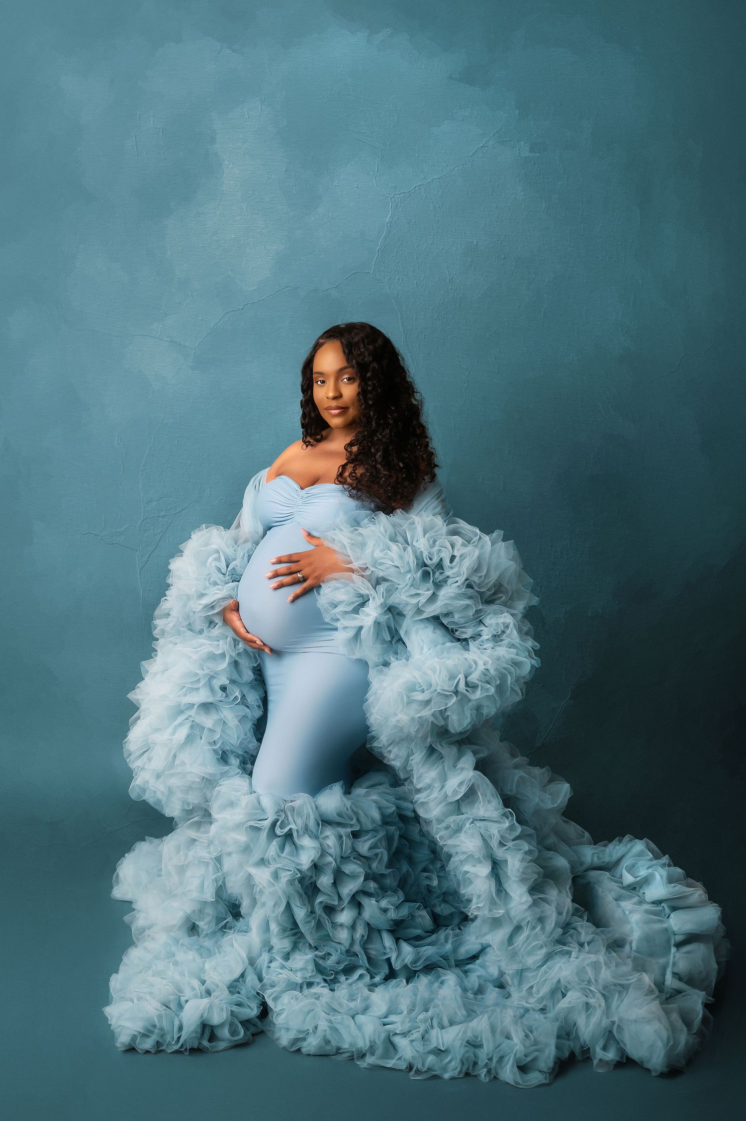Pregnant woman in a blue ruffle robe and fitted gown cradling her baby bump in a studio maternity photo.